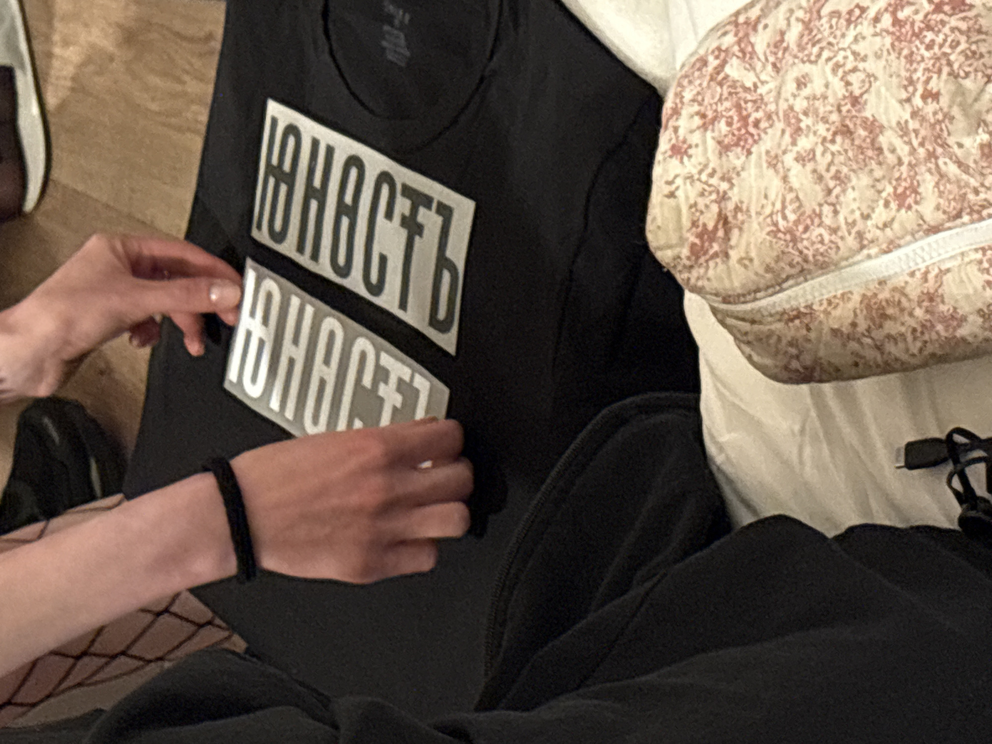 A person is applying a silkscreen print to a black T-shirt. The print is a word in Cyrillic script, possibly "Юность" (Youth). The scene occurs indoors and includes a floral-patterned pillow. The location is not explicitly stated within the visual content of the image, so Yerevan, Armenia, cannot be verified from the image alone.