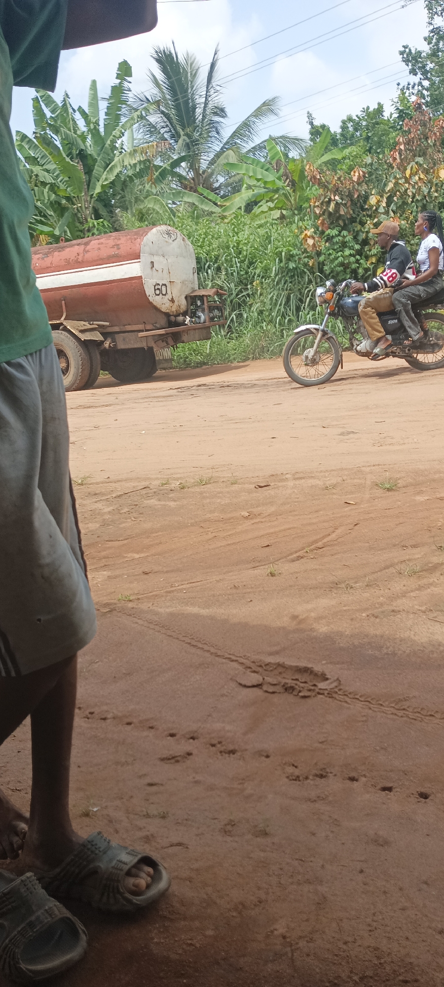 Amauzu, Nigeria: A dirt road scene. A brown and white tanker truck with the number "60" visible is parked on the roadside. A motorcycle with two people, a man driving and a woman riding as a passenger, passes the truck. Green foliage including banana trees line the background. In the foreground, a person wearing shorts and sandals is partially visible. The road is sandy with tracks. The sky is partially cloudy.