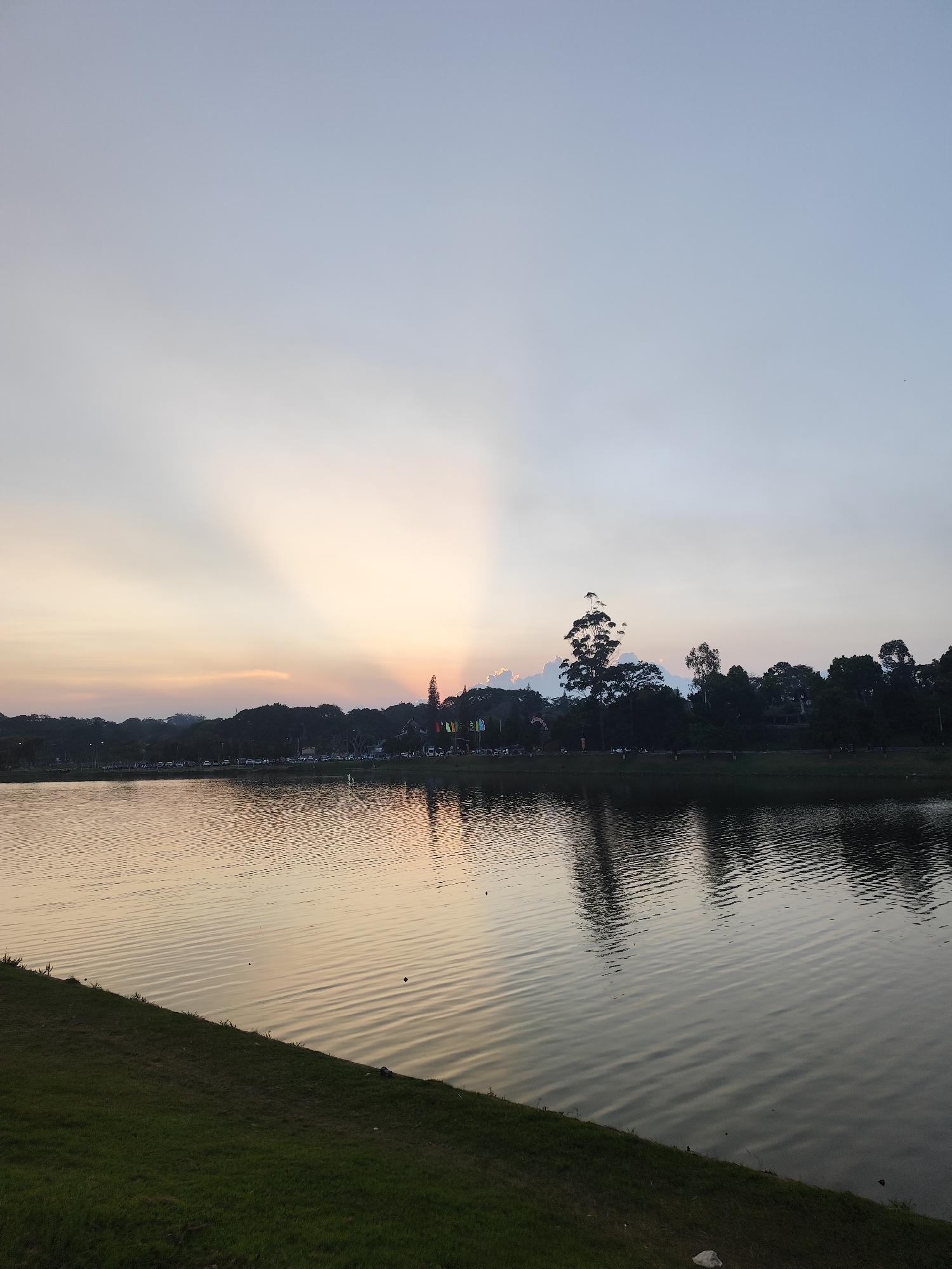 The image shows a body of water, likely a lake, with a grassy bank in the lower left corner. Across the water, a line of trees and some buildings are visible, with sunlight breaking through the clouds and trees behind. The sky is a mix of light blue and orange hues, suggesting sunset or sunrise. The water reflects the colors of the sky and the trees. Based on the general appearance, this is likely Xuan Huong Lake in Da Lat, Vietnam.