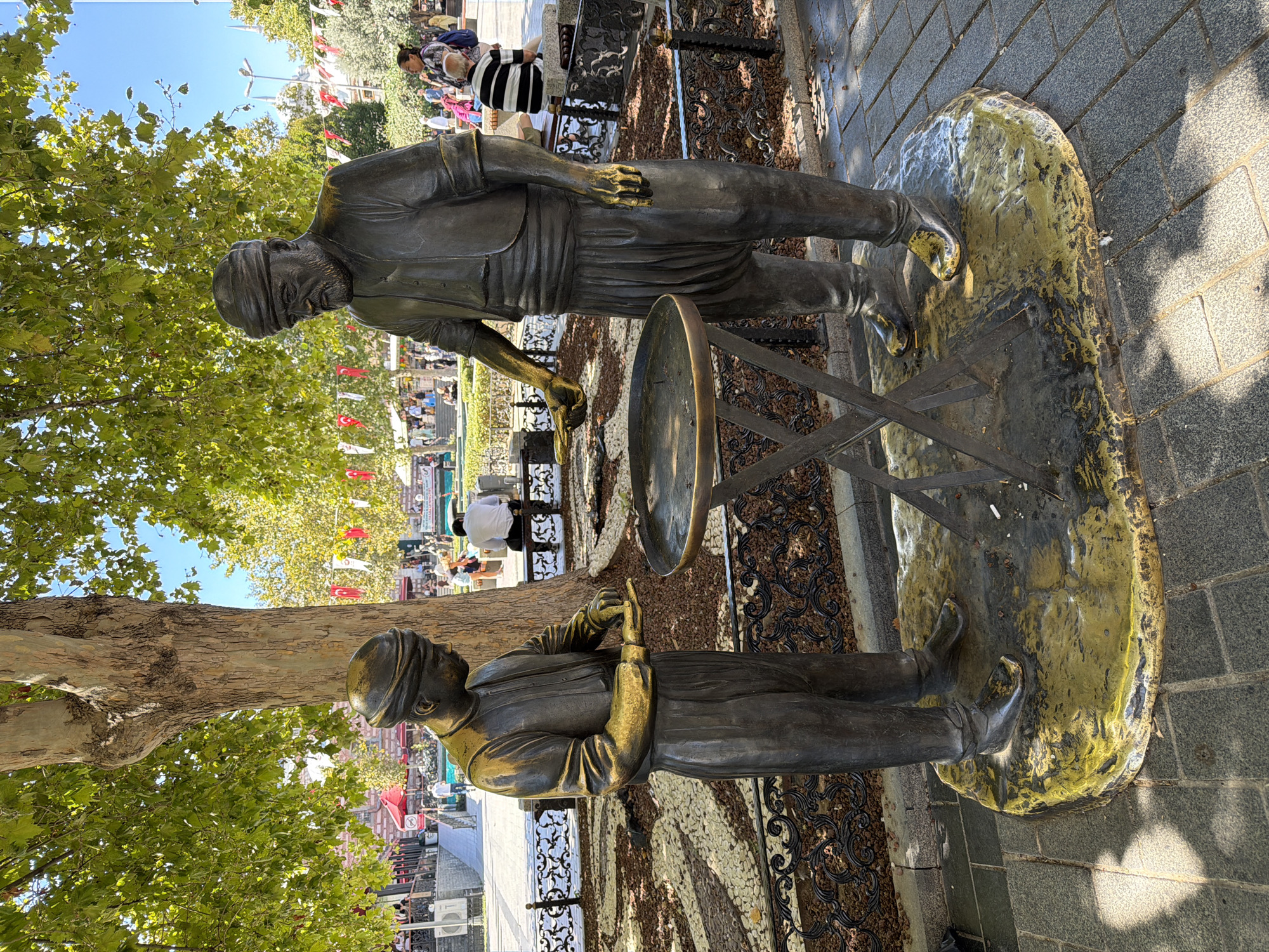 This is a photograph of a bronze statue depicting two men, likely street vendors, in an outdoor public space in Istanbul, Turkey. The statue is situated on a rough, yellow-gold painted base, surrounded by dark gravel and a decorative black metal fence. The larger statue on the right, an older man with a beard and turban, is looking down towards the smaller statue on the left. The younger man, also wearing a turban, is looking down at a round, shallow table between them, his hands positioned as if working with something on the table. Both figures are dressed in traditional Ottoman-era clothing, with the younger man wearing a vest and baggy trousers, and the older man wearing a tunic and similar trousers. The table appears to be a food cart or preparation surface.
The background shows a lively street scene with many people walking around, sitting on benches, and interacting. Buildings, trees, and decorative flags are visible. The overall impression is a bright, sunny day in a bustling city square. Based on the style of the statues, their attire, and the surrounding environment which suggests a historic or tourist area with stalls and public gathering spaces, this statue likely represents a traditional occupation that was once common in Istanbul, possibly depicting the preparation or sale of a local delicacy.