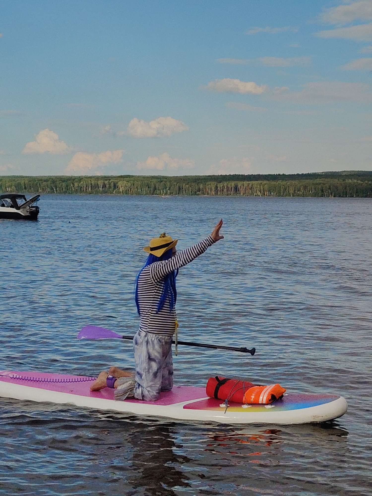 A person wearing a striped shirt, light-colored pants, a straw hat, and a blue wig kneels on a pink and white paddleboard. The person's right arm is raised with their hand extended. A purple paddle is resting on the paddleboard next to the person, and a black paddle shaft extends across the board. A red life vest and an orange safety vest are on the paddleboard near the front. The paddleboard is on a body of water with ripples. In the background, a line of trees forms the horizon under a blue sky with white clouds. A boat is visible in the far left background.