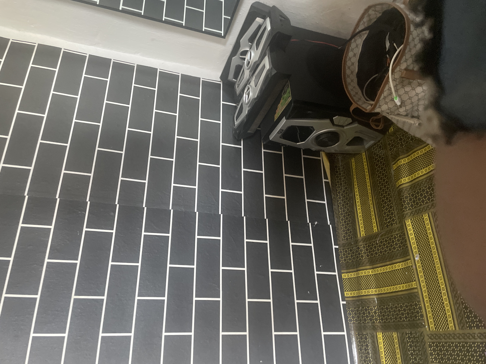The image shows a close-up view of a tiled floor and a variety of objects. The floor is covered with dark gray rectangular tiles arranged in a brick pattern, with white grout lines. To the right of the tiled floor is a patterned mat with yellow and dark green geometric designs.
On top of the mat and partially on the tiles, there are several objects. A black electronic device with silver accents and large speakers is visible. Next to it, a beige designer handbag with a repeating pattern is open, revealing dark contents and some charging cables. Part of a person's leg, clothed in dark fabric, is visible in the bottom right corner of the image.
The setting appears to be indoors, possibly a room with tiled flooring. The lighting suggests it could be daytime, but no specific time of day or weather conditions can be determined. There is no visible text in the image. The focus is on the textures and patterns of the floor, mat, and objects.