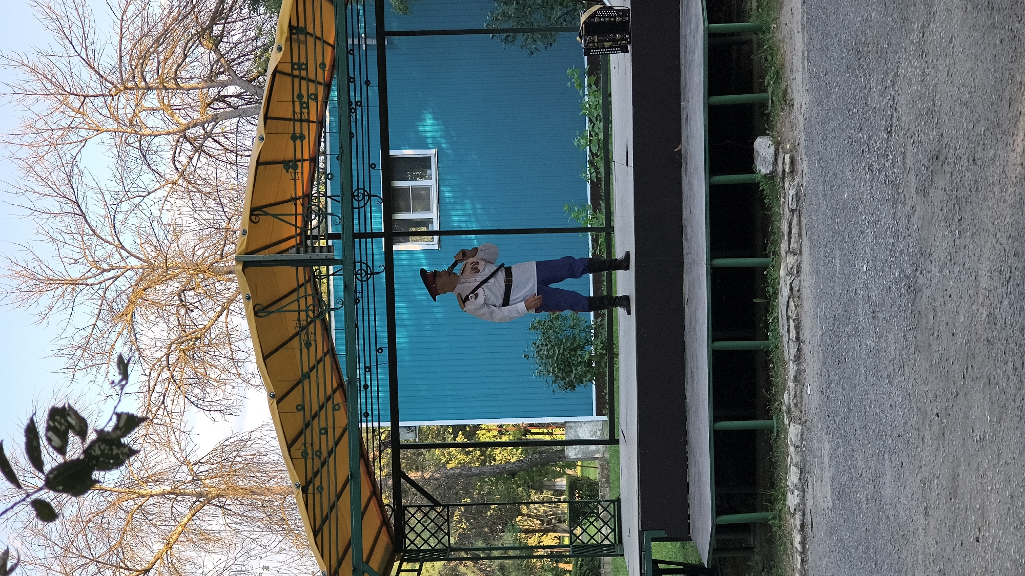 A lone male dressed in a traditional Cossack uniform, complete with a peaked cap, white shirt, dark belt, blue pants, and black boots, stands center stage on a raised platform. He is holding a microphone to his lips, appearing to sing or speak into it. Behind him, a bright turquoise building with a white-framed window serves as a backdrop. To the right of the stage, an accordion rests on the floor. The stage is covered by a yellow awning supported by a dark metal frame, situated outdoors under a clear sky with bare trees visible in the background. The overall setting suggests an outdoor performance or cultural event in Mikhaylovka, Russia.