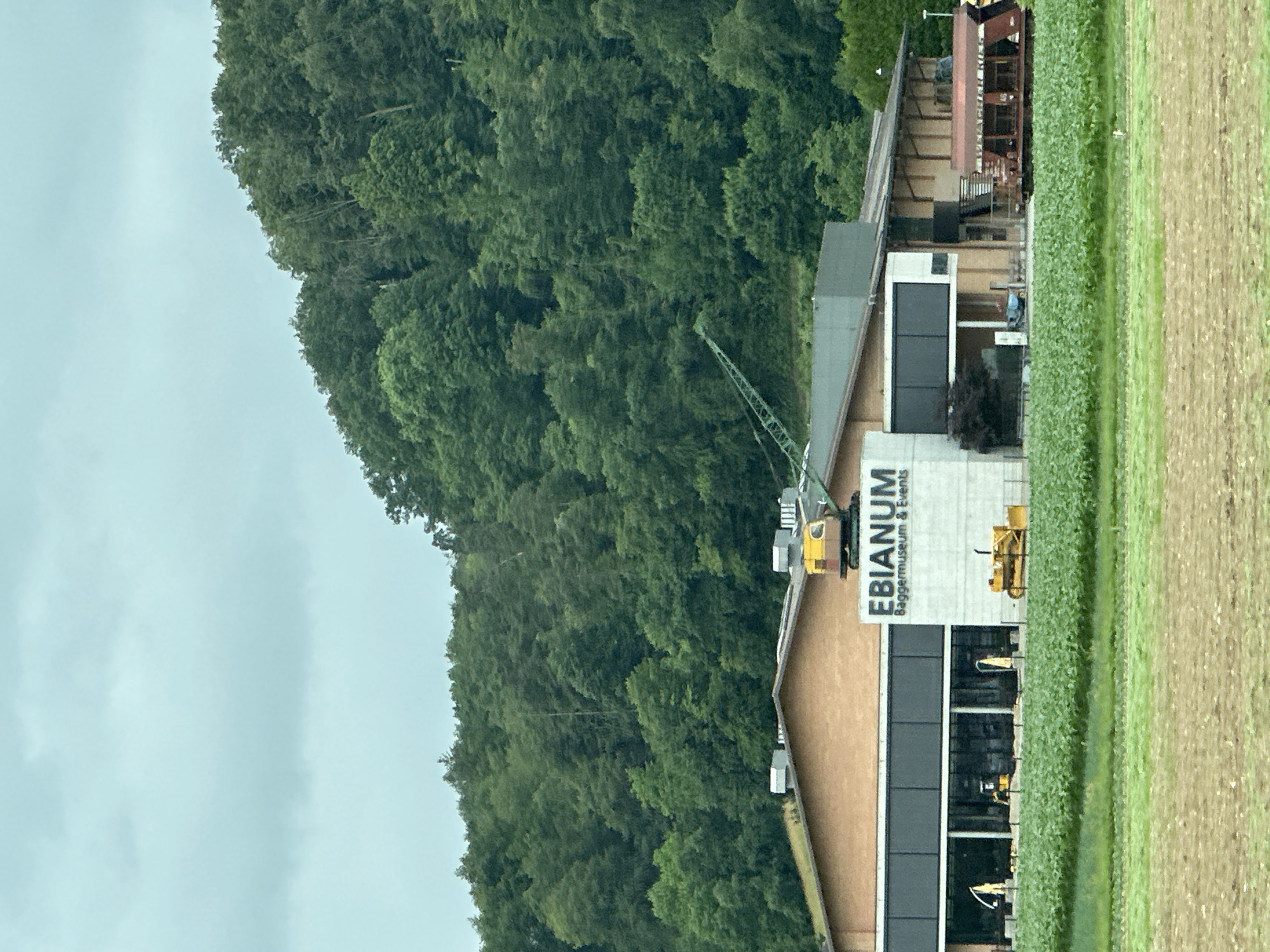 This image depicts the exterior of the EBianum Baggermuseum & Events facility in Zurzach, Switzerland. The building has a modern architectural design with a prominent white section displaying the museum's name. A large green crane is positioned on top of the building, and various construction vehicles, including yellow excavators, are visible in front of the facility. The museum is surrounded by lush green fields and a dense forest covering a hillside in the background. The sky is overcast, suggesting a cloudy day. The scene appears to be a static, daylight capture of the museum and its grounds.