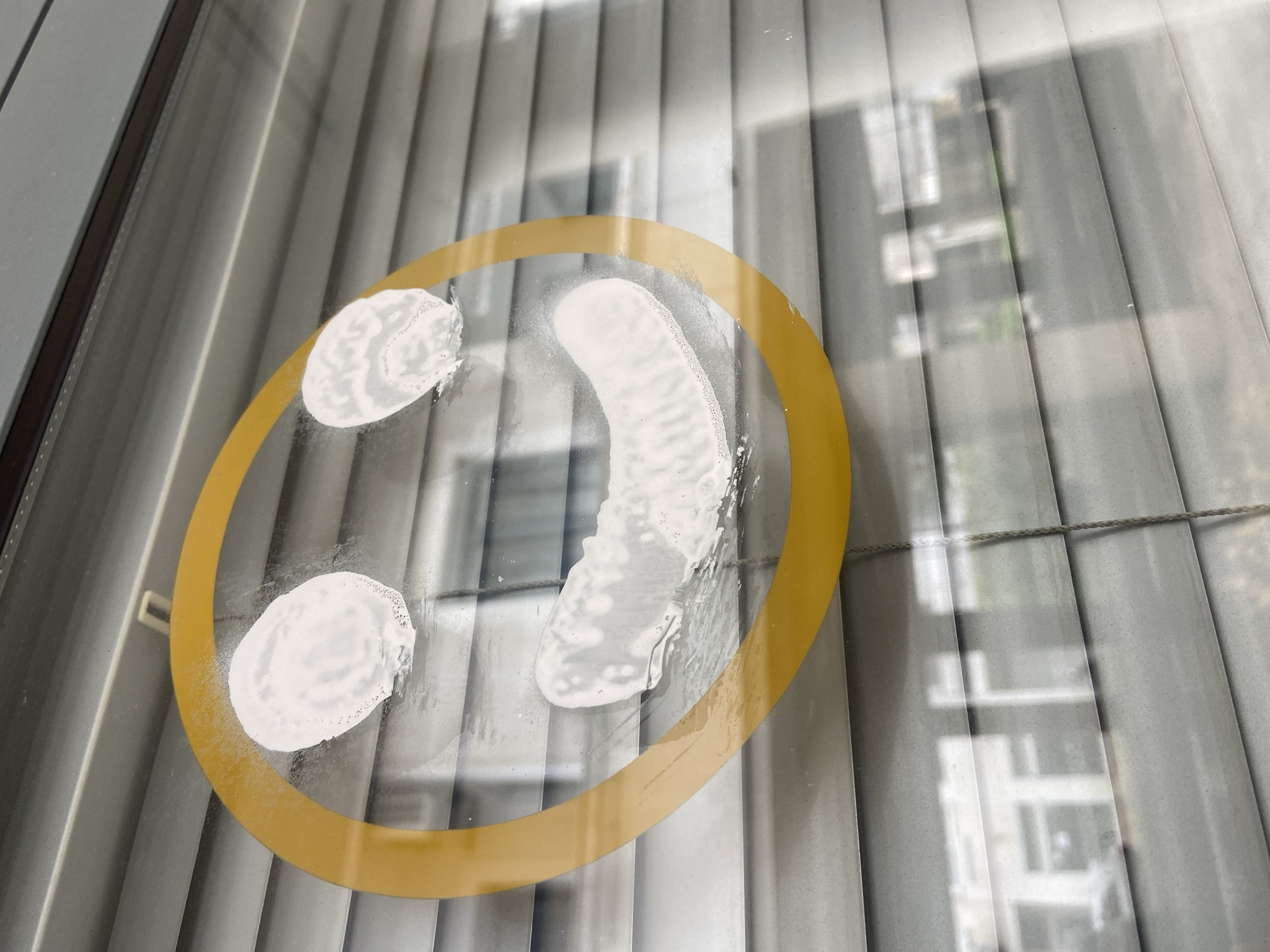 A close-up shot depicts a smiley face drawn on a glass surface, likely a window. The smiley face is created with a thick yellow outline and white, opaque fillings for the eyes and mouth. The background consists of vertical window blinds, and beyond them, a blurred view of a building exterior, giving the impression the window is an interior-facing one with a view of a structure outside. Based on the architectural style and environment visible in the blurred background, it is likely the location is in Budva, Montenegro, or a similar coastal area.