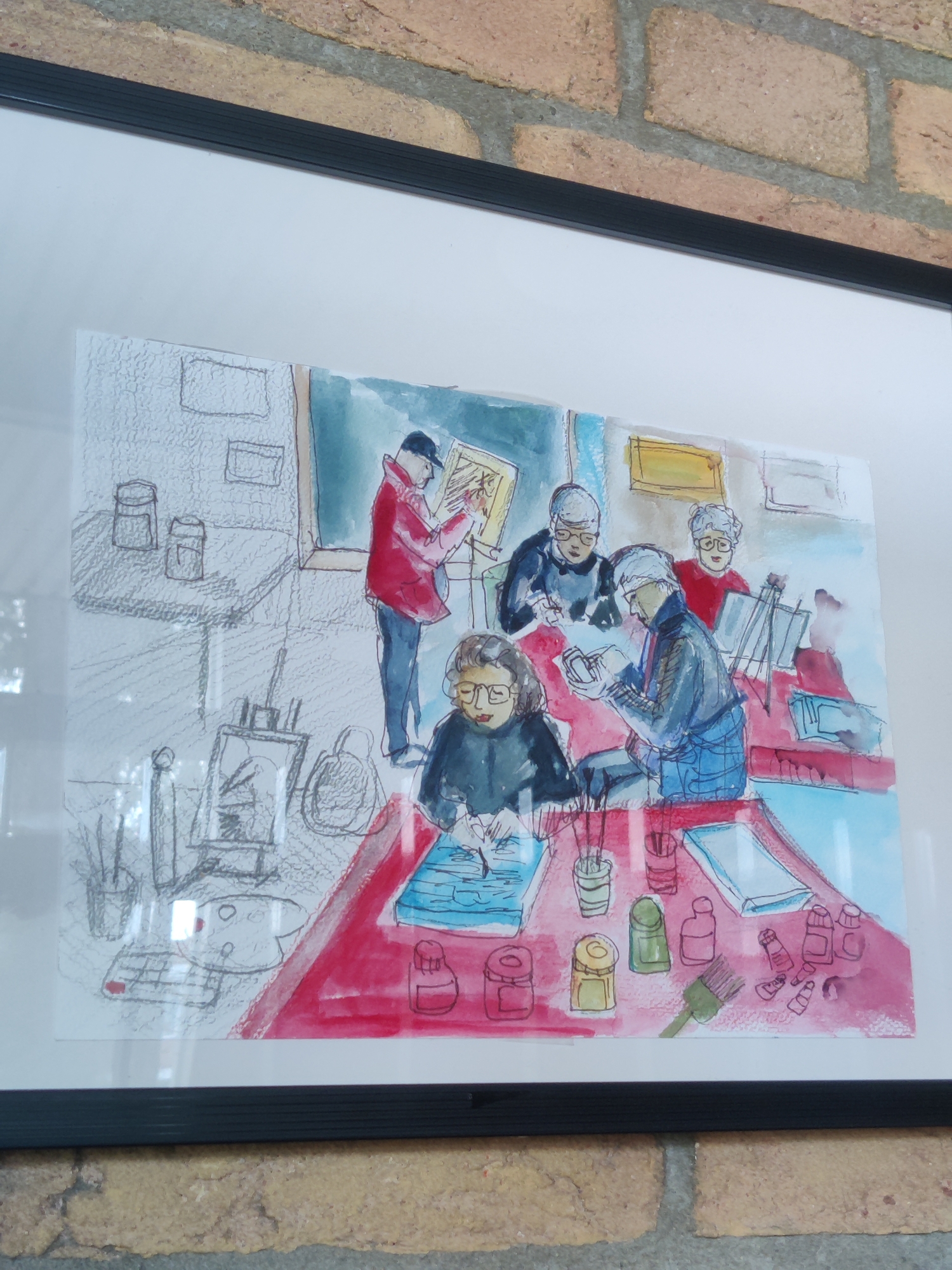 This is a framed watercolor and pencil sketch depicting an art class. Several individuals are seated around a long table, engaged in painting or drawing. One person stands, wearing a red jacket, and appears to be instructing or demonstrating. The room has a blackboard or large artwork in the background, and easels are set up. On the table, various art supplies are visible, including paint tubes, brushes, jars, and palettes. The overall style is loose and expressive, with a focus on capturing the dynamic of the group activity. The lighting suggests an indoor setting, and there are no specific cues for time of day or weather. The emotion conveyed is one of focused engagement and creative pursuit. There is no visible text in the artwork.