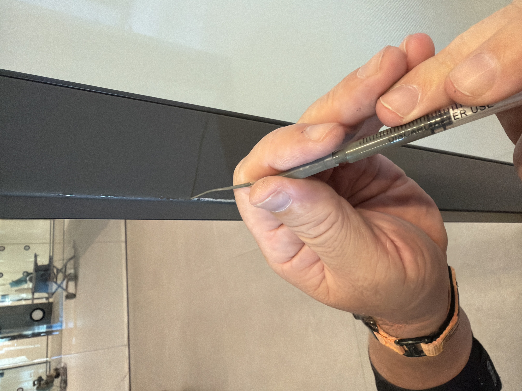 A close-up, eye-level shot captures a person's hands holding a syringe with a fine needle, poised to dispense a substance into a narrow seam on a dark gray vertical panel. The syringe, partially filled with a clear liquid, has black markings, including the words "ER USE ONLY". The person's left hand is visible on the wrist, wearing an orange and black watch. The background shows a brightly lit, modern interior with a reflective glass wall, offering a blurred glimpse of a small, wheeled cart and other elements, suggesting a public or commercial space. The overall scene is clean and well-lit, with the focus sharply on the hands and the syringe. The image does not provide any specific indicators of its location in Jerusalem, Israel.