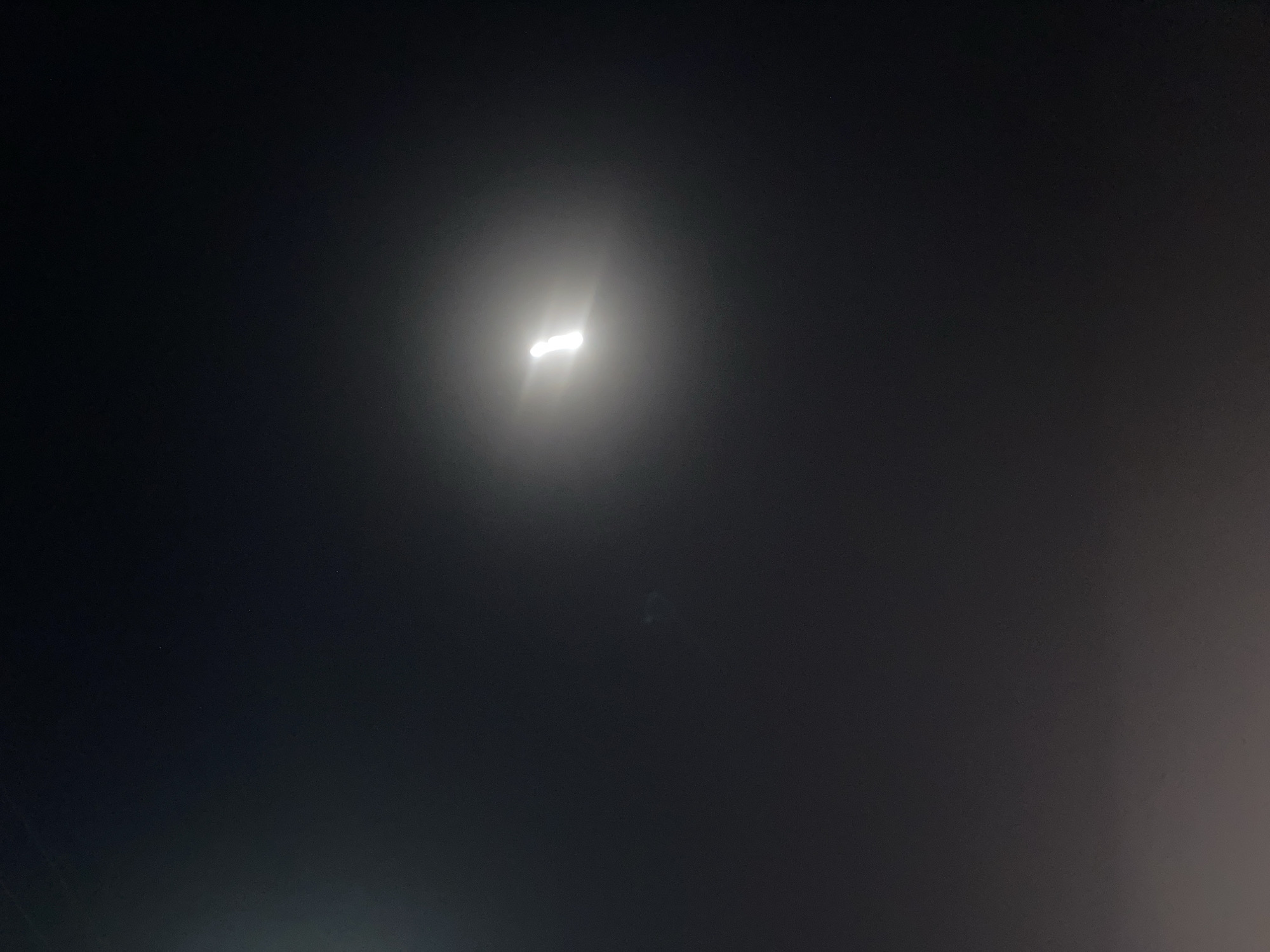 The image depicts a night scene in low visibility conditions, likely due to fog or haze. A bright light source is visible at the center of the frame, significantly diffused by the surrounding atmosphere. The overall tonality is dark, with gradients of gray indicating varying degrees of light diffusion. No people, specific subjects, or recognizable objects are discernible apart from the light. The environment is ambiguous due to the obscuring effect of the fog. The location in the city of Unknown, Unknown cannot be confirmed.