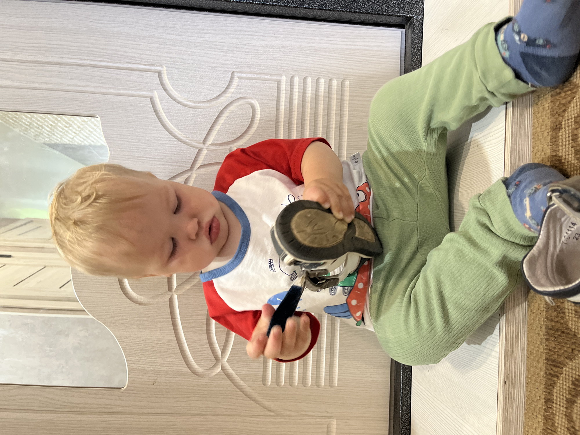 A fair-haired toddler sits on the floor in front of a light-colored wooden door with carved detailing. The child wears a red and white raglan t-shirt with a blue collar and a graphic print, light green ribbed pants, and blue socks with a car pattern. The child is holding a grey and white shoe in their hands, examining it closely. The shoe appears to be a child's sneaker. The flooring is a light wood-grain laminate, and a small section of a brown rug is visible in the foreground. The scene is likely indoors, possibly a home or a children's facility, within the city of Chaikovsky, Russia.