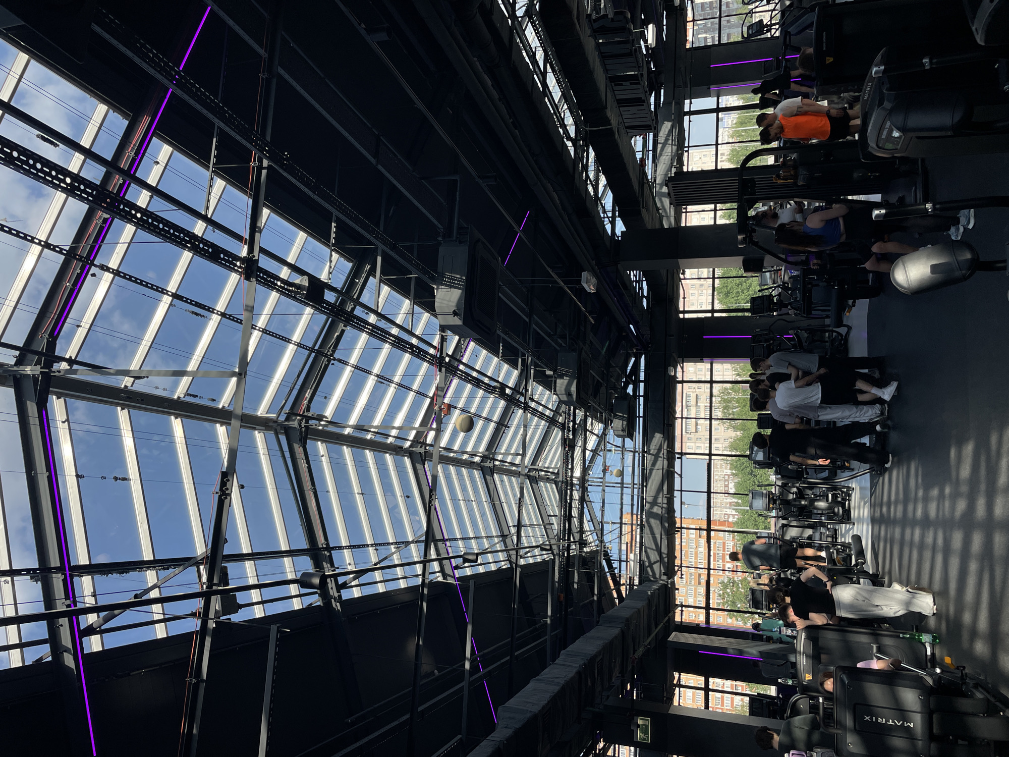 The image shows the interior of a gym in Kazan, Russia. The location is identifiable by buildings seen outside through large windows. People are using exercise equipment, including seated machines. A distinctive architectural feature is a large glass roof supported by a black metal framework. Purple neon lights are visible on some of the supporting structures.
