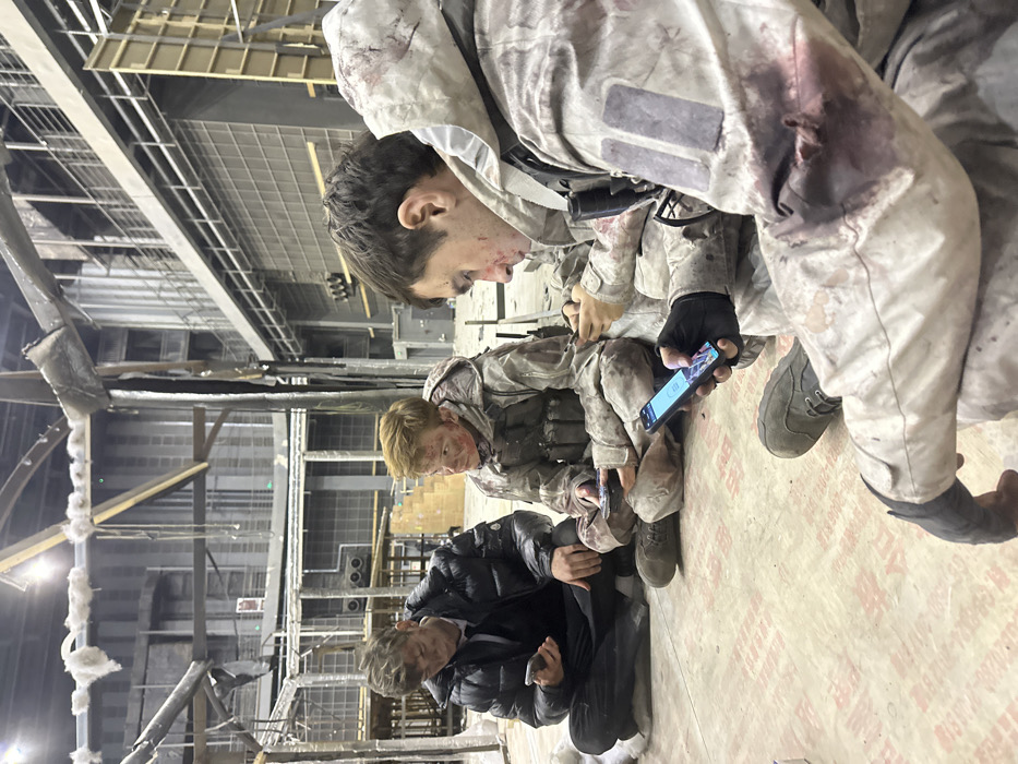 The image displays three male individuals, appearing to be actors, seated on a light-colored floor within a large, industrial-style film soundstage or production facility located in Huangdao District, China. The background reveals a complex set construction featuring exposed steel beams, scaffolding, and various structural elements suggesting a damaged or war-torn environment, with insulation material hanging from overhead components. Overhead artificial lighting is visible.
All three individuals are dressed in military-style or tactical clothing, heavily stained with simulated dirt, grime, and fake blood, consistent with costume and makeup effects. The leftmost individual wears a black puffer jacket over dark clothing, with a dirt-smeared face, and is looking down at a smartphone in his hands. The middle individual is clad in a light-colored camouflage uniform with a visible tactical vest and pouches, his face showing smudges of dirt and simulated blood; he is also focused on a smartphone. The rightmost individual, positioned in the foreground, wears a light-colored, possibly hooded, combat uniform with prominent simulated bloodstains on his right arm and chest. He has simulated blood on his face, wears a black glove on his right hand, and is actively engaging with a dark-colored smartphone, whose screen displays an application with multiple icons. A black tactical flashlight is clipped to his uniform near the shoulder. All three individuals are independently engaged with their respective smartphones, indicating a break or period of inactivity on the set.