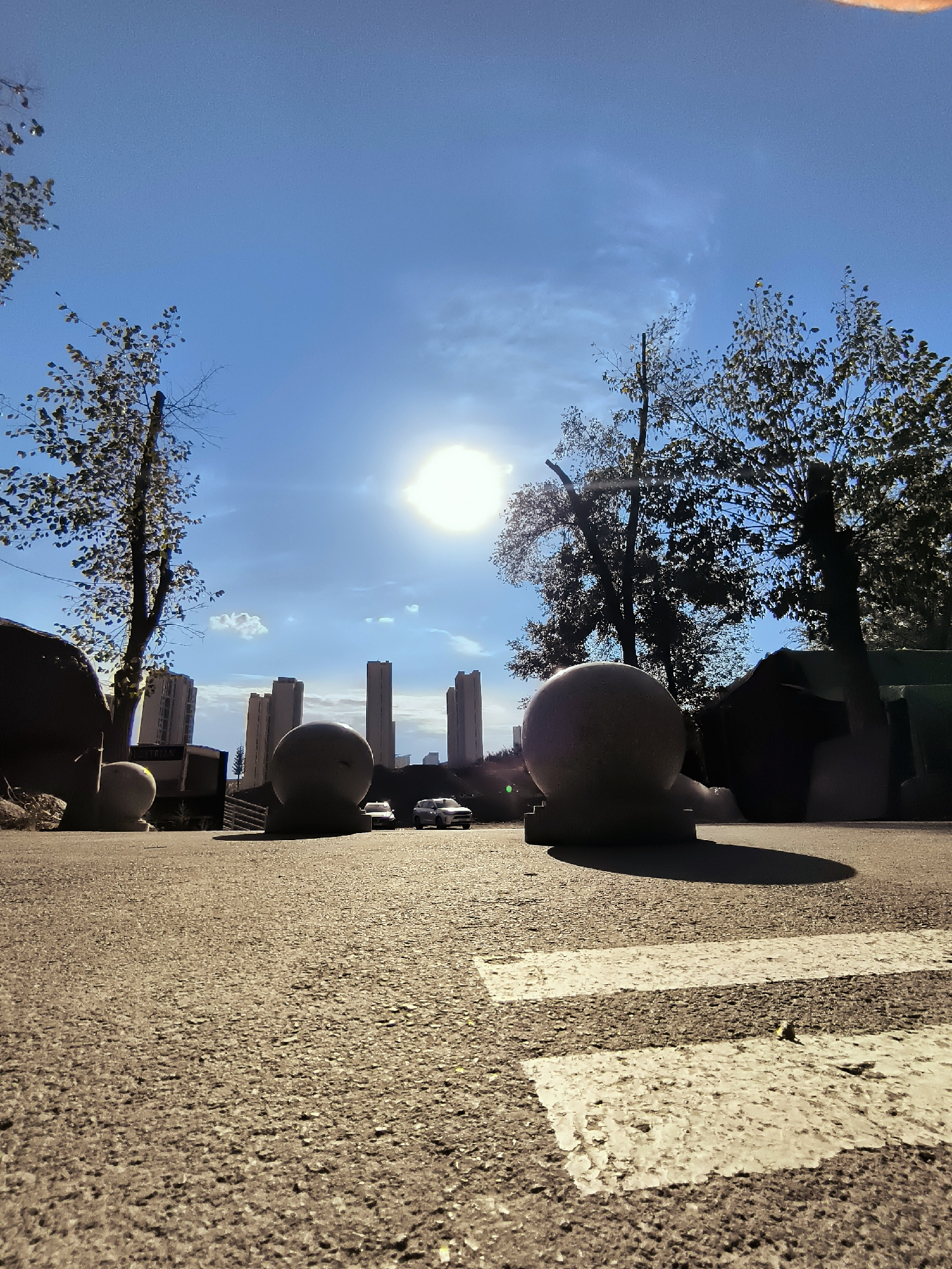 The low-angle, wide-shot photograph captures a paved area in Ürümqi, China, with a bright sun in the sky casting long shadows. In the foreground, a pedestrian crossing with white stripes is visible on the asphalt. Two large, spherical concrete bollards are prominent. One is to the left, partially obscured by what appears to be a large rock. The other is centrally positioned to the right, casting a distinct shadow. Behind the bollards, two white SUVs are parked. In the mid-ground, several modern, tall buildings rise against the blue sky. Trees with sparse foliage are scattered throughout the scene, silhouetted against the bright sky on the left and right. The overall impression is of an urban public space bathed in sunlight.