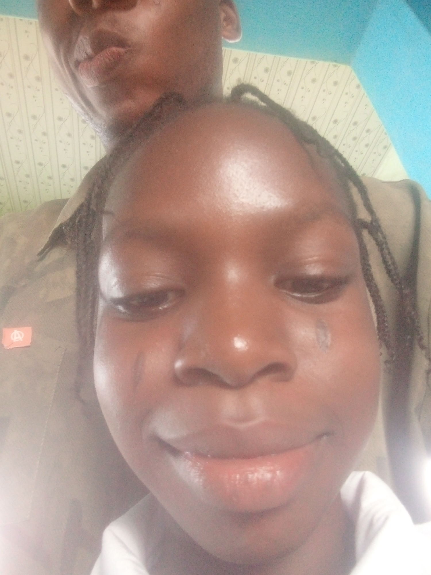 The image shows two individuals. The primary subject is a young, dark-skinned person with braided hair looking down. A streak is visible on their left cheek. The second person, appearing in the background, is an adult with dark skin. The background is partially visible, revealing a blue wall and a patterned wallpaper. The location of Ikorodu, Nigeria is not identifiable based on the image content.