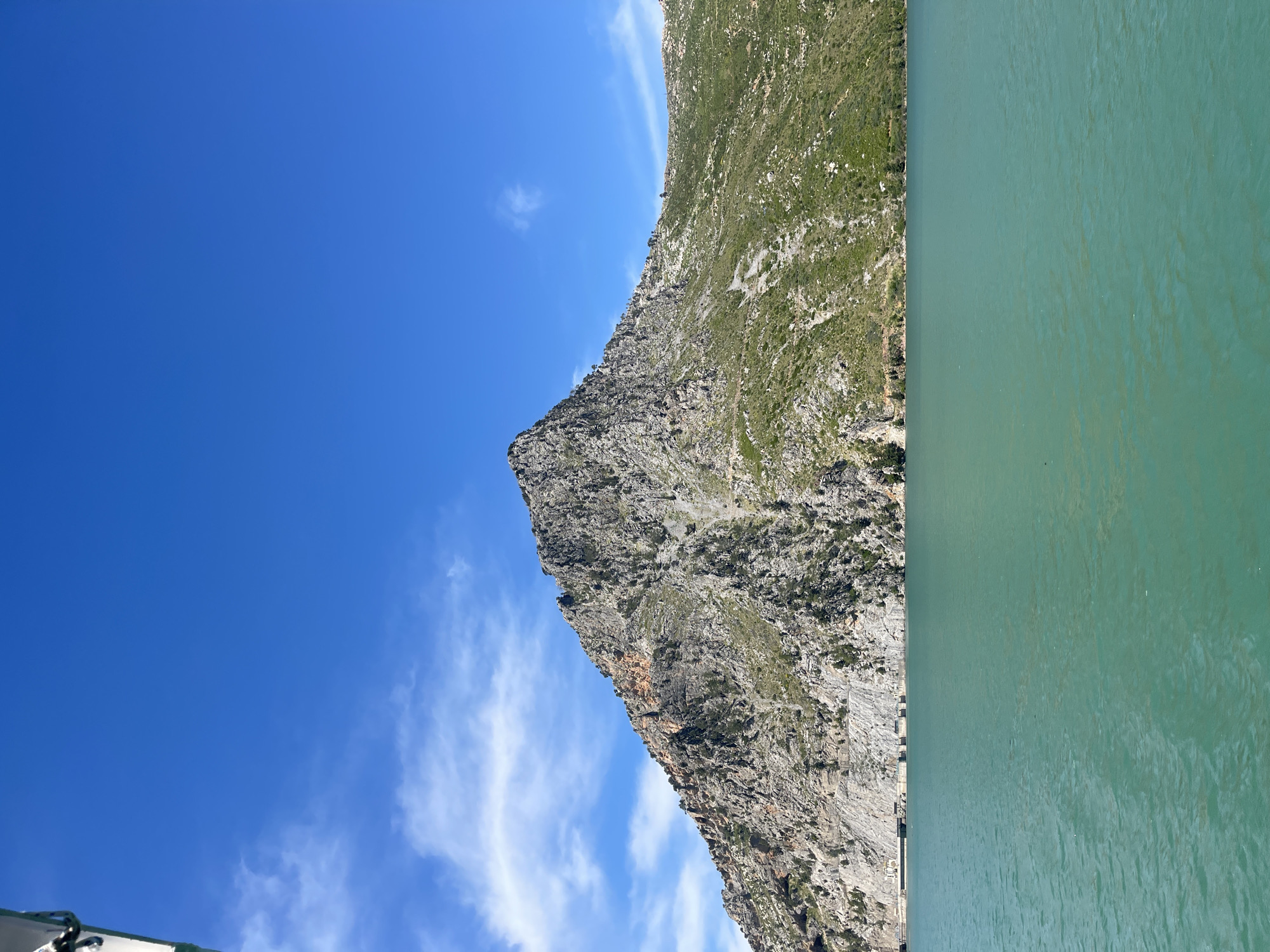 A landscape photo shows a mountain adjacent to a body of water, with a blue sky above. The mountain is rocky with some green vegetation on its slopes. The body of water is green-blue. The sky is clear with a few wispy clouds. The photo is rotated 90 degrees counter-clockwise. The location is Unknown, Unknown.