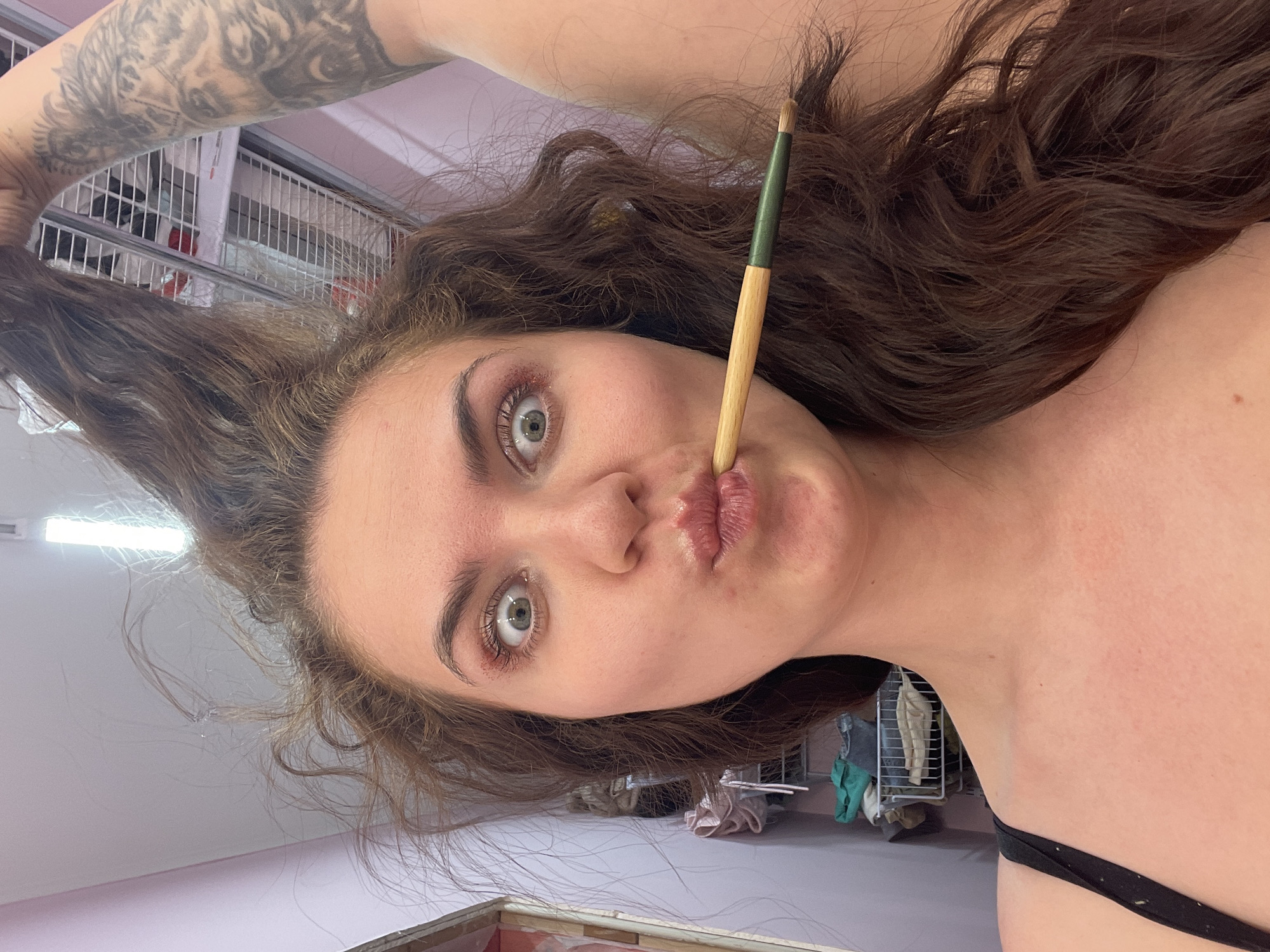 A fair-skinned woman with brown, curly hair is holding a paintbrush in her mouth and pulling a section of her hair upward. She has gray-green eyes and pink lips. Her left arm is raised, revealing a tattoo. The background appears to be an indoor setting with a white ceiling, metal shelving units, and various items stored on them. There is no evidence to directly place the image in Мосметаллоконструкция Онт, Russia.