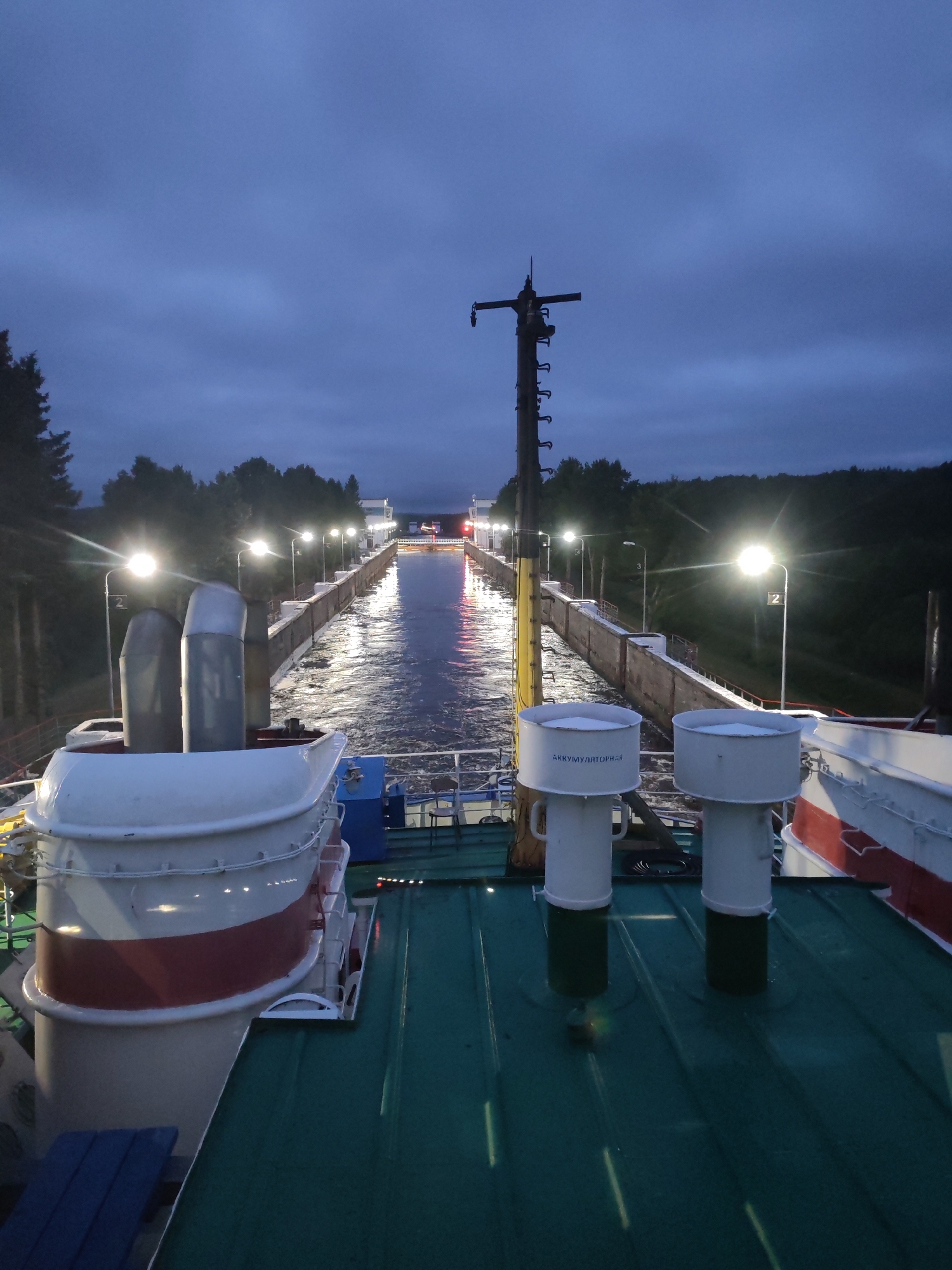 The image depicts a view from the deck of a ship, looking forward into a canal lock at dusk. The sky is dark blue with some clouds. The canal is lined with concrete walls and illuminated by rows of lights on poles. The water in the canal is rippling and reflects the lights. On the deck of the ship in the foreground, there are two white cylindrical structures with red stripes on their lower halves. These appear to be life rafts. Further back, there are two more white cylindrical structures, likely vents or exhaust pipes. A tall wooden pole with various attachments stands prominently on the right side of the canal. Trees line the canal banks on either side. The overall scene suggests a ship navigating through a lock system in a somewhat remote, possibly rural, location in Russia, given the text on one of the cylindrical structures which appears to be in Cyrillic. The location in Markovo, Russia, is not visually ascertainable from the image itself.