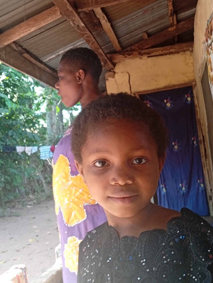 A young girl with short curly hair and a black, sequined top is centered in the frame. Behind her, slightly out of focus, stands an older individual with short hair, wearing a purple and yellow patterned garment. The background features a weathered building with a rustic wooden porch and a blue floral curtain. Laundry lines with hanging clothes are visible in the distance through the trees.