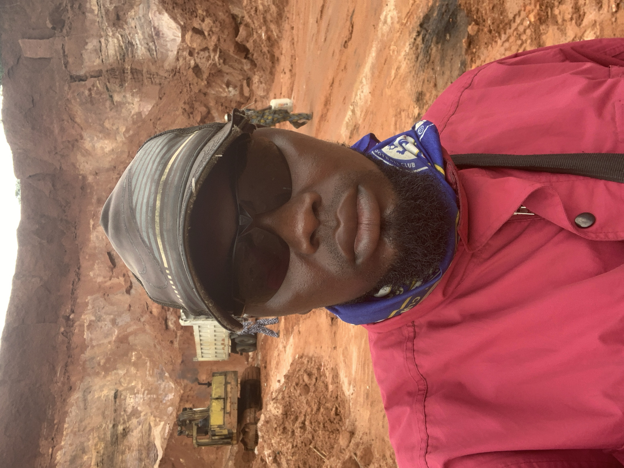The image shows a close-up selfie of a man wearing a dark baseball cap, sunglasses, and a red jacket. He has a beard and a blue bandana around his neck. In the background, there is a large excavation site with red earth. A yellow excavator and a white dump truck are visible, along with other workers in the distance. The scene appears to be a construction or mining site in Ajeregun, Nigeria, indicated by the red earth and the presence of heavy machinery. The lighting suggests it is daytime.