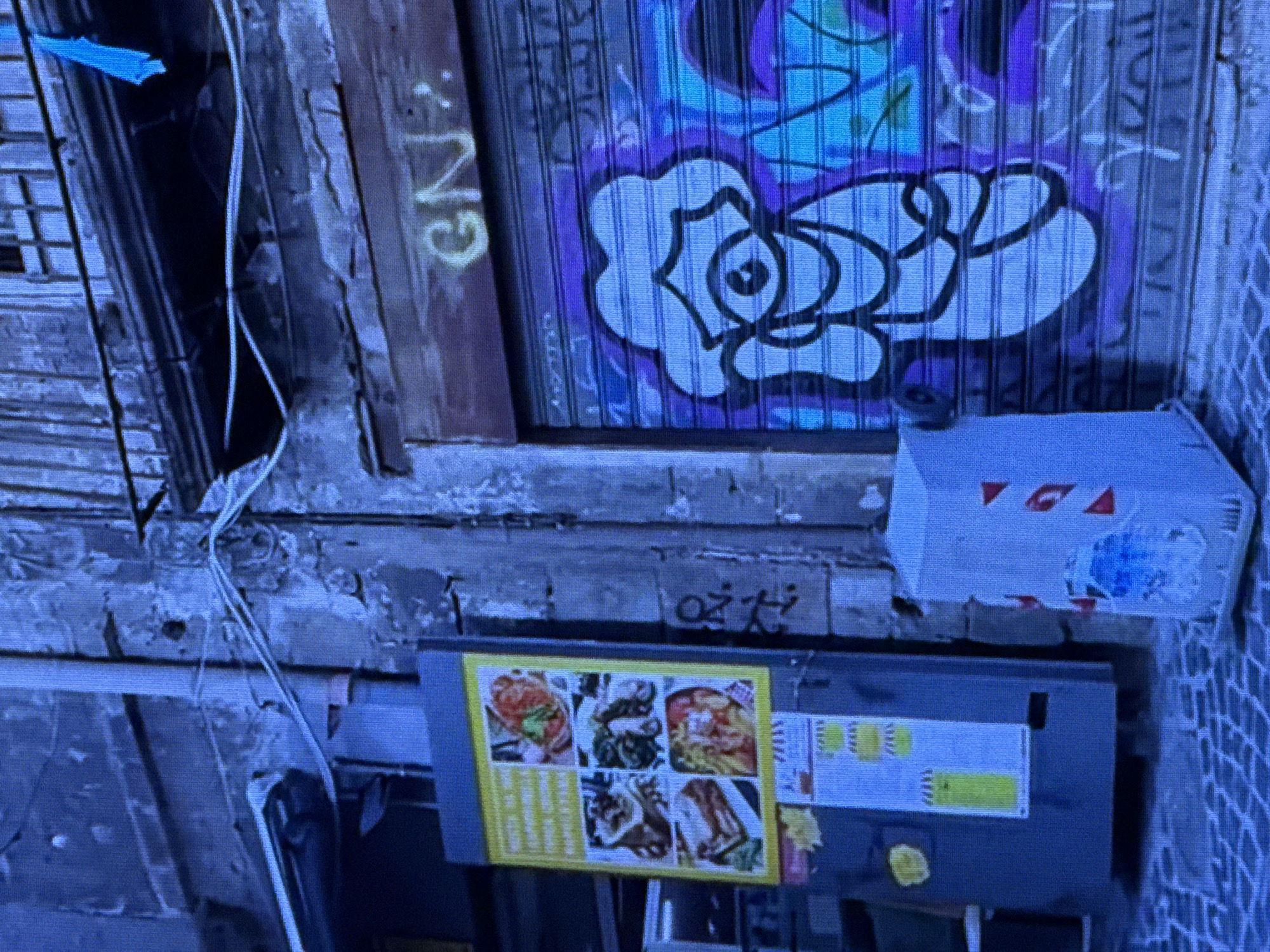 The image depicts an outdoor scene in a city, characterized by a textured, aged building facade with visible signs of wear and tear. Several objects are present, including a vertical advertising display featuring images of food and text, a small, cylindrical trash receptacle adorned with red triangular stickers, and a large, metallic roll-up door covered in graffiti. The graffiti on the roll-up door is colorful and features a cartoonish character with a large eye and abstract shapes, along with some legible text. The ground appears to be paved with cobblestones. The overall lighting and color palette suggest it might be evening or nighttime, with a strong blue cast to the image. There are no people visible in the scene.