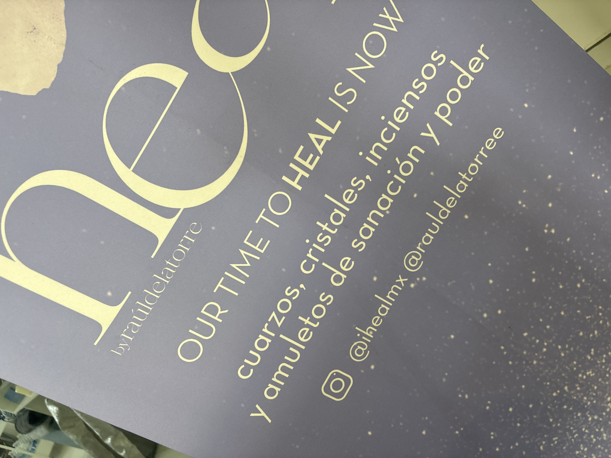 The image displays a close-up, angled view of a vertical, light-purple banner. Prominent stylized pale-yellow lettering at the top partially shows "he," followed by "by raúldelatorre." Below this, the English phrase "OUR TIME TO HEAL IS NOW" is rendered in pale-yellow uppercase letters. Further down, the Spanish text "cuarzos, cristales, inciensos y amuletos de sanación y poder" (meaning "quartz, crystals, incense, and healing and power amulets") is visible. The bottom section of the banner includes an Instagram logo alongside the handles "@ihealmx @rauldelatorree." The banner's surface is speckled with numerous small, pale-yellow dots. A minor, irregular brown smudge or tear is present near the top right edge. The background is out of focus, indicating an indoor setting with blurred white and light-colored shelving or display units. No individuals or specific actions are visible. The branding implies a Mexican context, but the image lacks explicit details to determine a precise location within the city of Puebla.