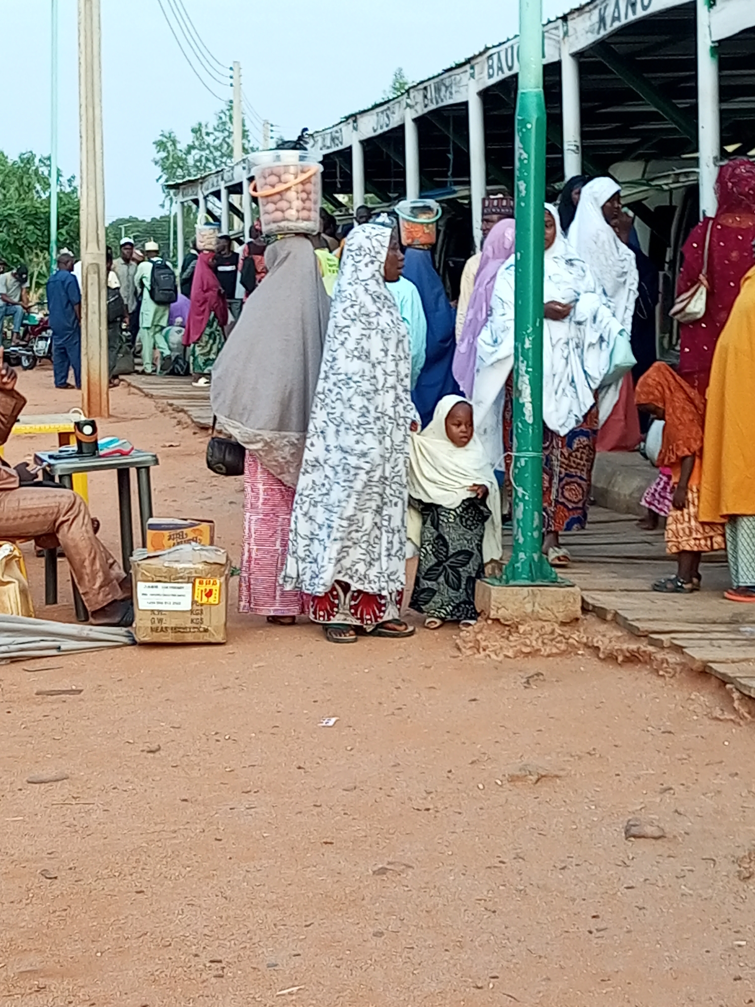 The image depicts a busy scene at what appears to be a bus station in Gombe, Nigeria. Several individuals, primarily women and children, are gathered, some carrying goods. In the foreground, a person is seated by a small table with a speaker and what looks like shoes. Boxes are stacked near the table. A woman in the center is carrying a bucket filled with eggs on her head, a common sight in markets. Various individuals are dressed in traditional Islamic attire, with headscarves and flowing garments. The background shows a structure with signage that partially reads "BAU KAN...". The ground is sandy and dusty, typical of the environment. The overall impression is one of everyday commerce and travel.