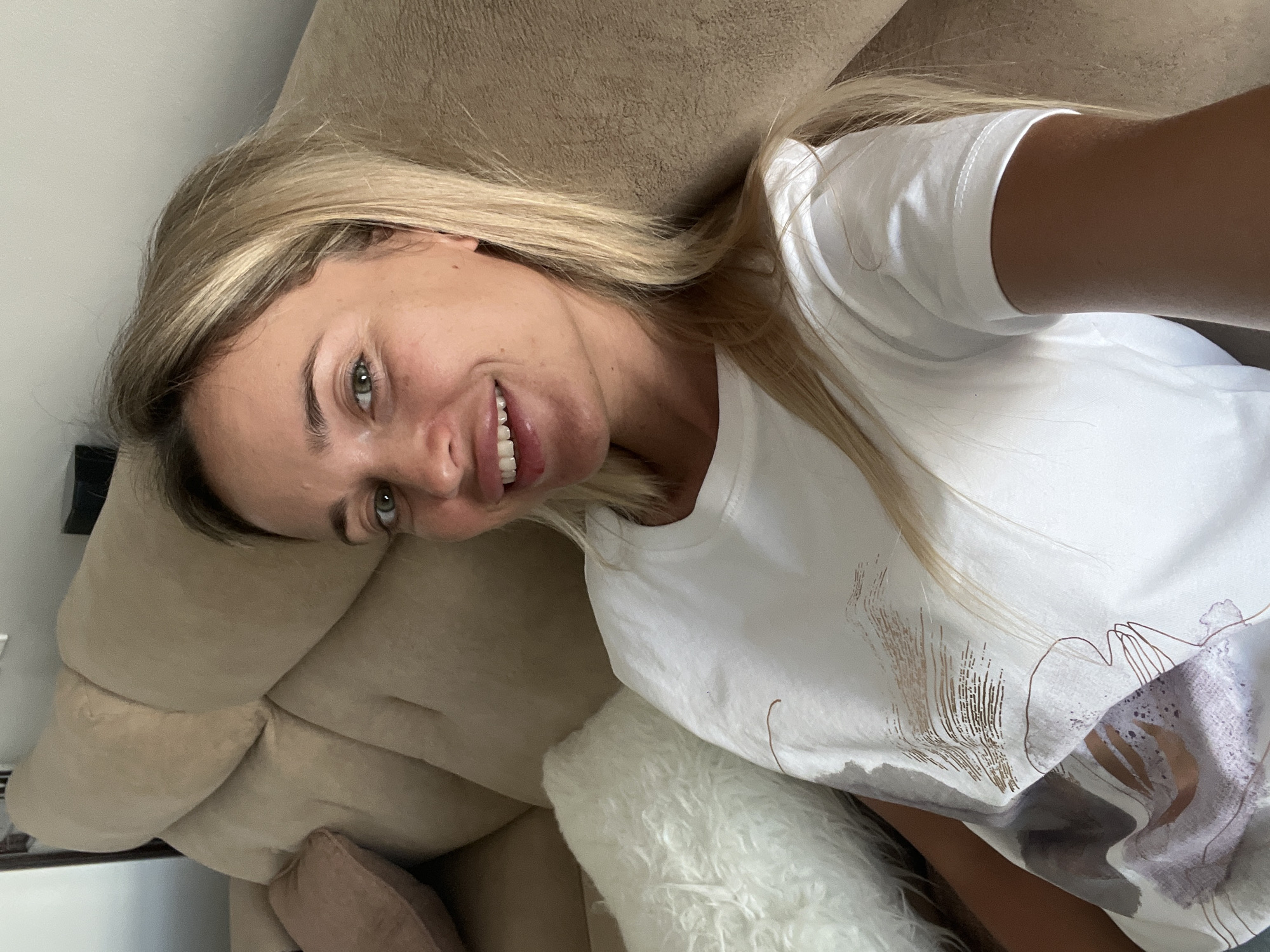 A woman with blonde hair is sitting on a sofa. She is wearing a white t-shirt with a printed design. She is smiling and looking at the camera. The sofa is a light brown color and has several cushions. There is a white fluffy cushion next to the woman. The background is a wall. The image does not provide any specific details about the city of Sharm El Sheikh, Egypt.