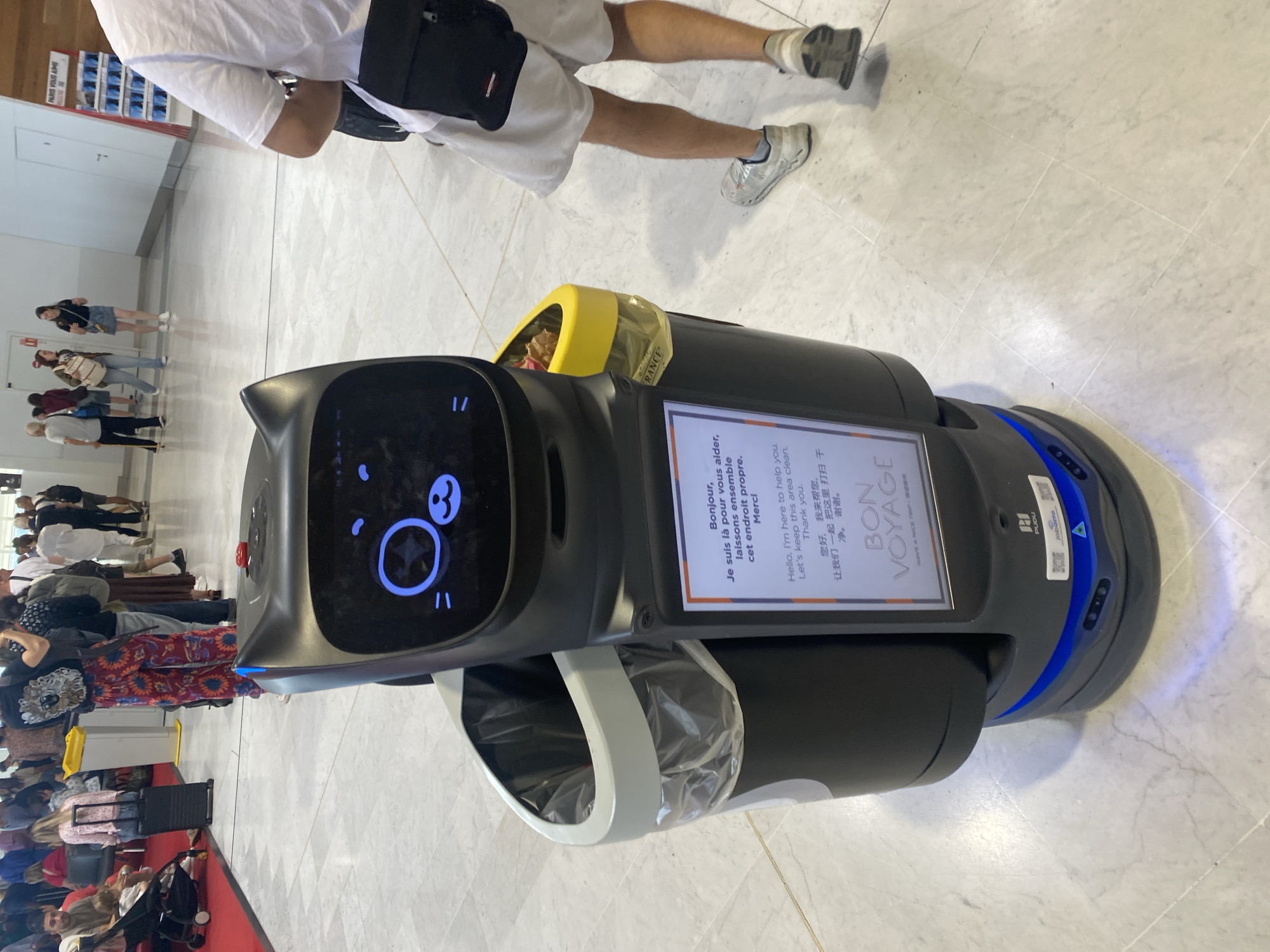 In the city of Tremblay-en-France, France, a robotic trash can with a cat-like screen is positioned in a well-lit indoor area, possibly an airport terminal, with a marble tiled floor. The robot has a black body, a white rim around its top opening, and a yellow compartment for trash. A screen on its front displays a message in English, French, and Chinese: "Bonjour, Je suis là pour vous aider, Laissons ensemble cet endroit propre. Merci. Hello, I'm here to help you. Let's keep this area clean. Thank you. 您好,我来帮忙,让我们一起把这里打扫干净,谢谢." Below this, the text "BON VOYAGE" is visible. In the background, a line of people, some with luggage, are waiting. A person in white shorts and a white t-shirt is walking away from the camera to the right of the robot.