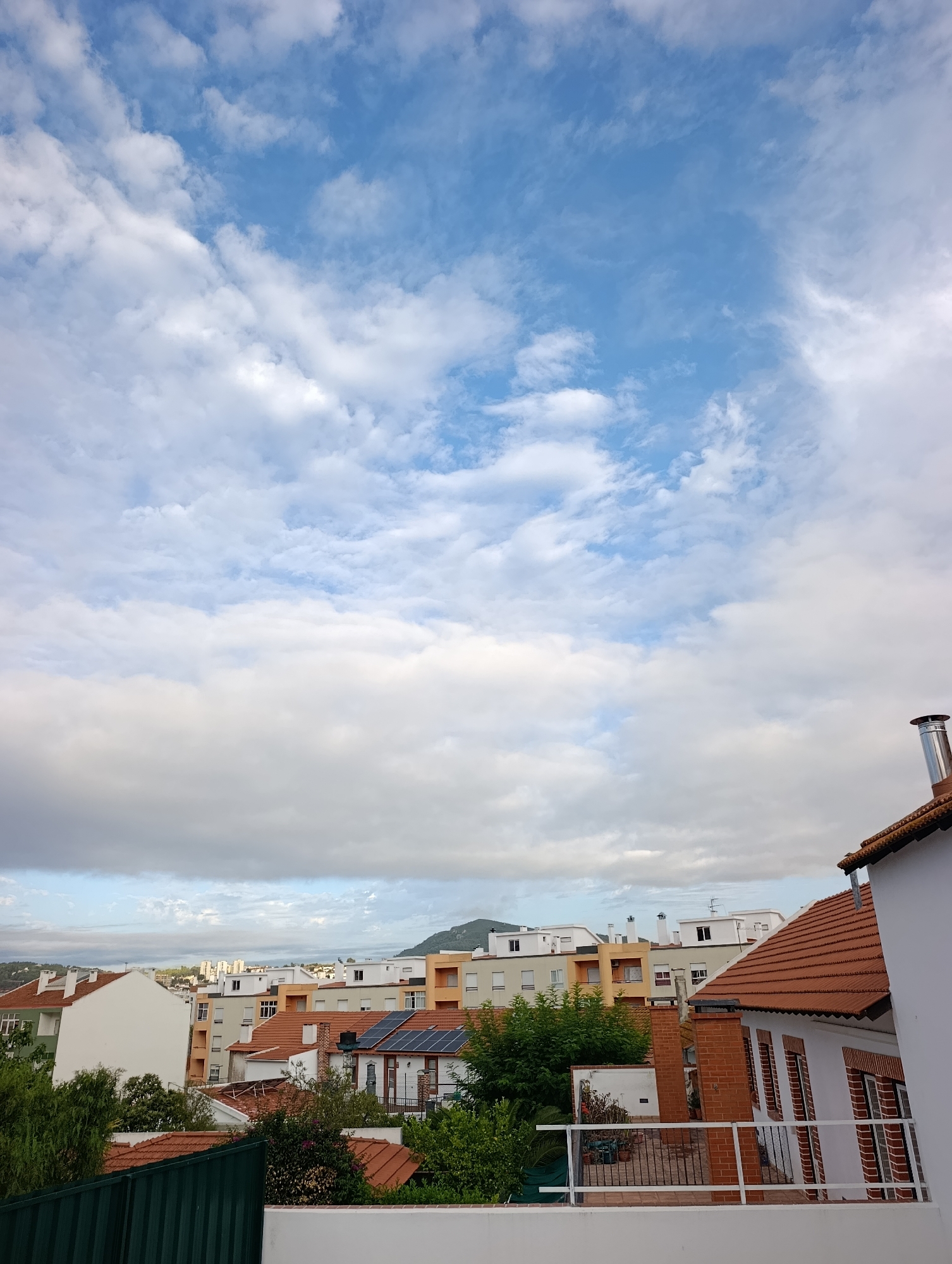 The image shows a view of a residential area in Setúbal, Portugal, under a partly cloudy sky. The foreground is dominated by the roof and side of a white house with a terracotta tiled roof. A metal chimney protrudes from the roof of a brick structure connected to the house. A white railing encloses a balcony area.

Beyond this house, a collection of buildings can be seen. Many have terracotta tiled roofs. Some buildings are taller apartment blocks with white, beige, and orange facades. Solar panels are visible on the roof of one of the houses. Green trees and foliage are interspersed throughout the scene. In the distance, more buildings and a hilly landscape are visible under a sky filled with blue patches and white clouds, some of which are darker and appear closer. The overall impression is of a sunny day with some cloud cover in a Portuguese city.