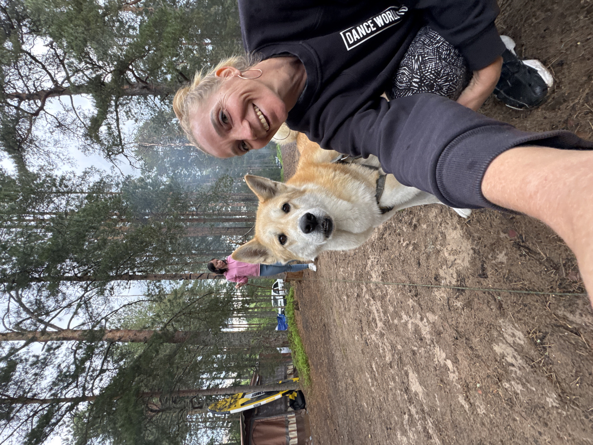 An outdoor selfie photograph captured in a wooded area in Zelenaya Roshcha, Russia, depicts two women and a dog. The primary subject, a blonde woman with her hair in a bun, occupies the right side of the frame. She is smiling broadly, looking directly at the camera, and wearing a black long-sleeved sweatshirt with "DANCE WORLD" printed in white, along with dark leggings featuring a white geometric pattern. She wears gold hoop earrings. Her right arm is extended, holding the camera, while she crouches on the muddy ground.
Adjacent to the main subject, slightly left of center, stands a medium-sized dog with light brown and white fur, wearing a dark harness. The dog is looking directly at the camera with a neutral expression. A thin green leash extends from its harness towards the left.
In the mid-ground, a second woman with dark hair stands wearing a pink hooded sweatshirt and light blue jeans. She appears to be holding a thin line or rope in her right hand and is looking towards the left, away from the camera.
The environment consists of a dense forest of tall coniferous trees under an overcast sky. The ground is reddish-brown earth, appearing damp and marked with paths, with sparse vegetation. On the far left, a yellow and black stand-up paddleboard or surfboard leans against a brown, tent-like structure. Further in the background, a white car is partially visible, parked near a blue tarp on the ground. Wisps of smoke rise from behind the trees in the mid-ground, suggesting a campfire or cooking activity.