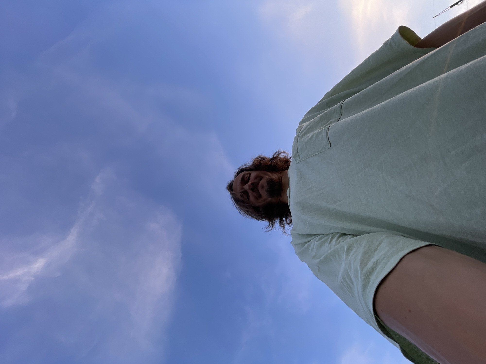 An overhead, low-angle, close-up shot of a man with medium-length, wavy brown hair and a goatee is captured against a clear blue sky with scattered wispy clouds. He is wearing a light green t-shirt with a breast pocket. The man is looking directly at the camera with a slight smile. His right arm is visible in the foreground, bent at the elbow, and his left arm extends out of frame. The background consists entirely of the sky, with a hint of a sunset glow on the right side. The scene appears to be outdoors.
