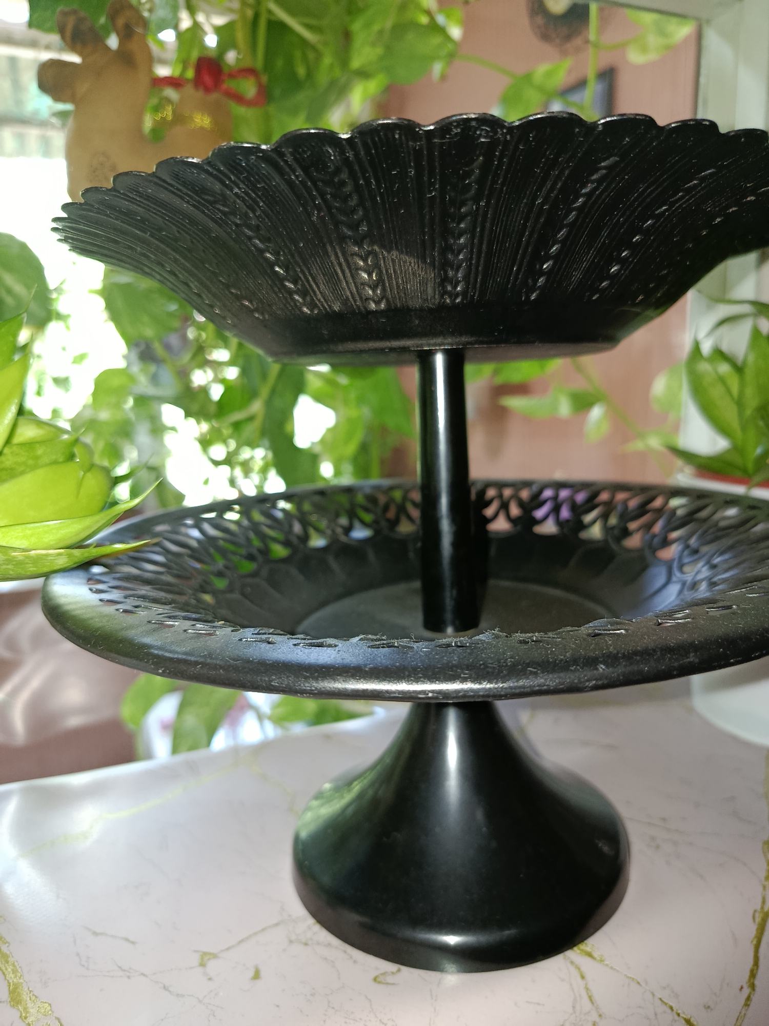 The image displays a close-up, eye-level view of a black, two-tiered serving stand. The stand is made of a matte material and features decorative edges on both tiers. The top tier has a fluted design with a pattern resembling stylized leaves or grains, while the bottom tier has an open, filigree-like pattern. A central black pillar connects the two tiers, and the stand rests on a dark, flared base. The stand is positioned on a light-colored surface with a marble-like pattern, featuring subtle green and yellow veining. In the background, out of focus, are lush green plants, suggesting an indoor setting with natural light. A small, light brown, deer-shaped ornament with a red ribbon is partially visible hanging from the plants above the top tier. The overall impression is of a decorative household item, likely used for serving food, placed within a home environment. There is no indication of the specific location in Tashkent, Uzbekistan.