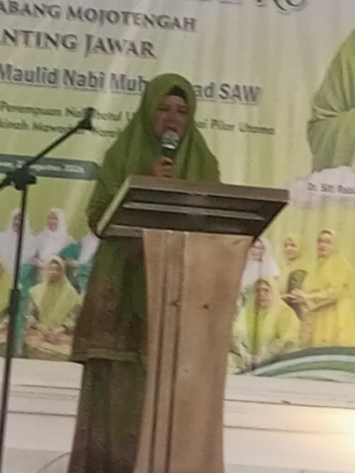 A woman wearing a green hijab stands behind a wooden podium and speaks into a microphone. She is in the foreground of the image. Behind her, a banner with text is visible. The text on the banner, partially obscured by the woman, appears to be in Indonesian and mentions "Maulid Nabi Muhammad SAW." The banner also has an image of people wearing hijabs. The event appears to be taking place indoors, possibly at a community hall or religious center in Klaseman, Indonesia, indicated by the banner's text.