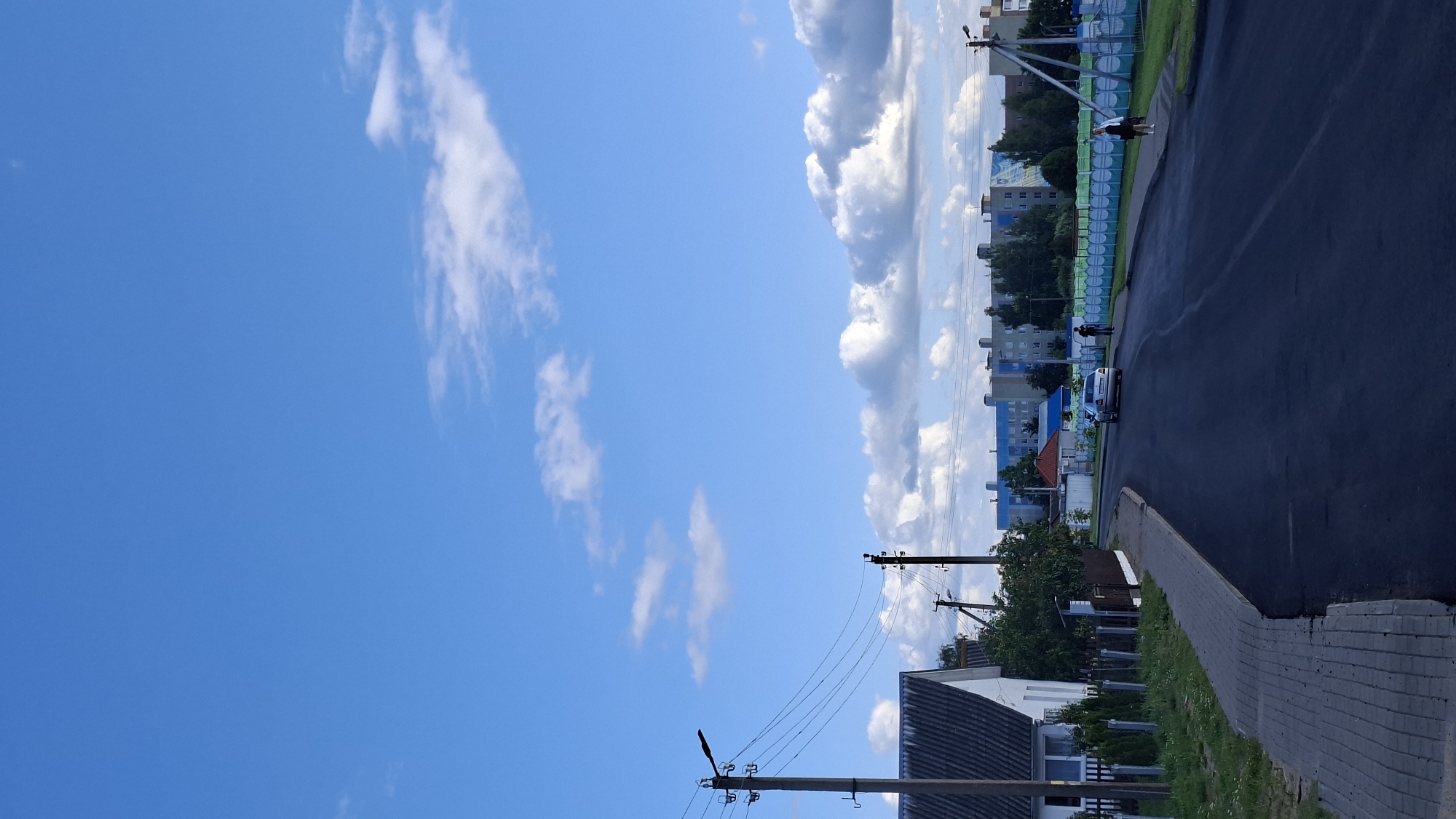 The image depicts a street scene in Myadzyel, Belarus, likely taken on a bright, sunny day with scattered clouds. The sky is a vibrant blue, filling the left side of the frame. On the right, an asphalt road runs parallel to a paved sidewalk and a grassy verge. Several residential buildings and apartment blocks are visible in the background, with some featuring blue and green decorative panels. Trees and utility poles with overhead wires are interspersed throughout the scene.
A silver car is parked on the side of the road. Two individuals are standing on the sidewalk near the car, and another person is further down the street, appearing to walk away from the camera. The overall atmosphere suggests a typical daytime scene in a residential area. There is no visible text in the image.