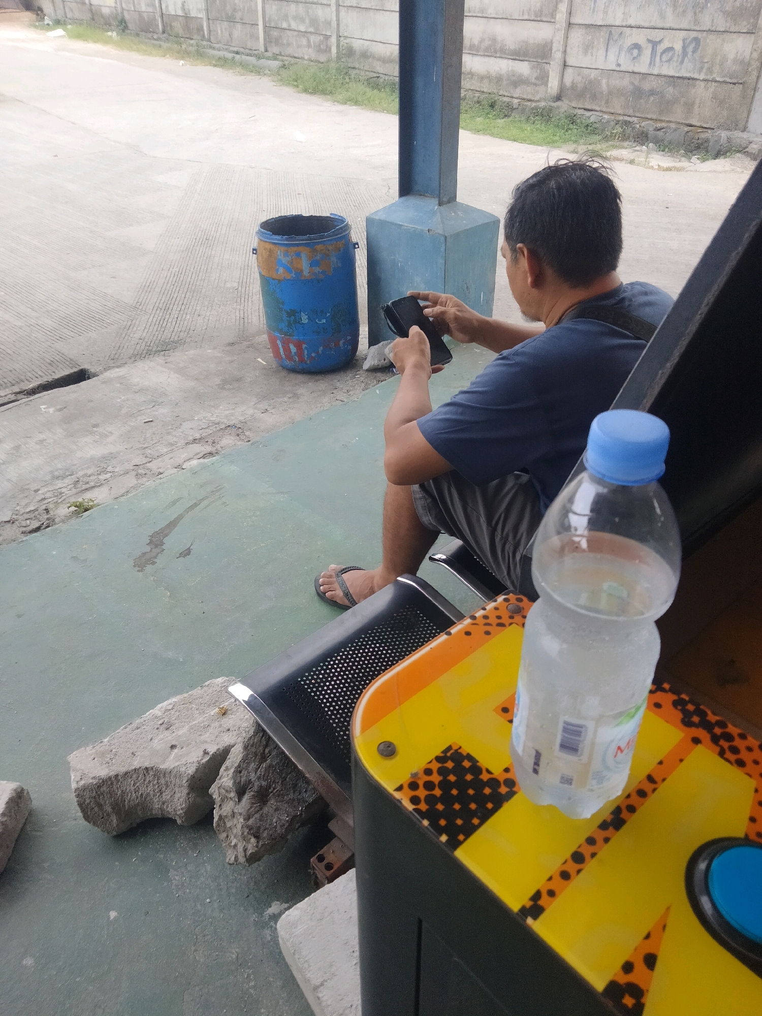 In Sukaasih, Indonesia, a man sits on a black perforated bench, holding a smartphone. He is wearing a dark blue t-shirt and grey shorts, and has dark hair. A blue-capped bottle of water sits on a yellow and black patterned surface in the foreground. Behind the man, a blue and orange drum barrel stands on a concrete surface, next to a concrete wall with graffiti. The environment appears to be outdoors, possibly a waiting area or public space.