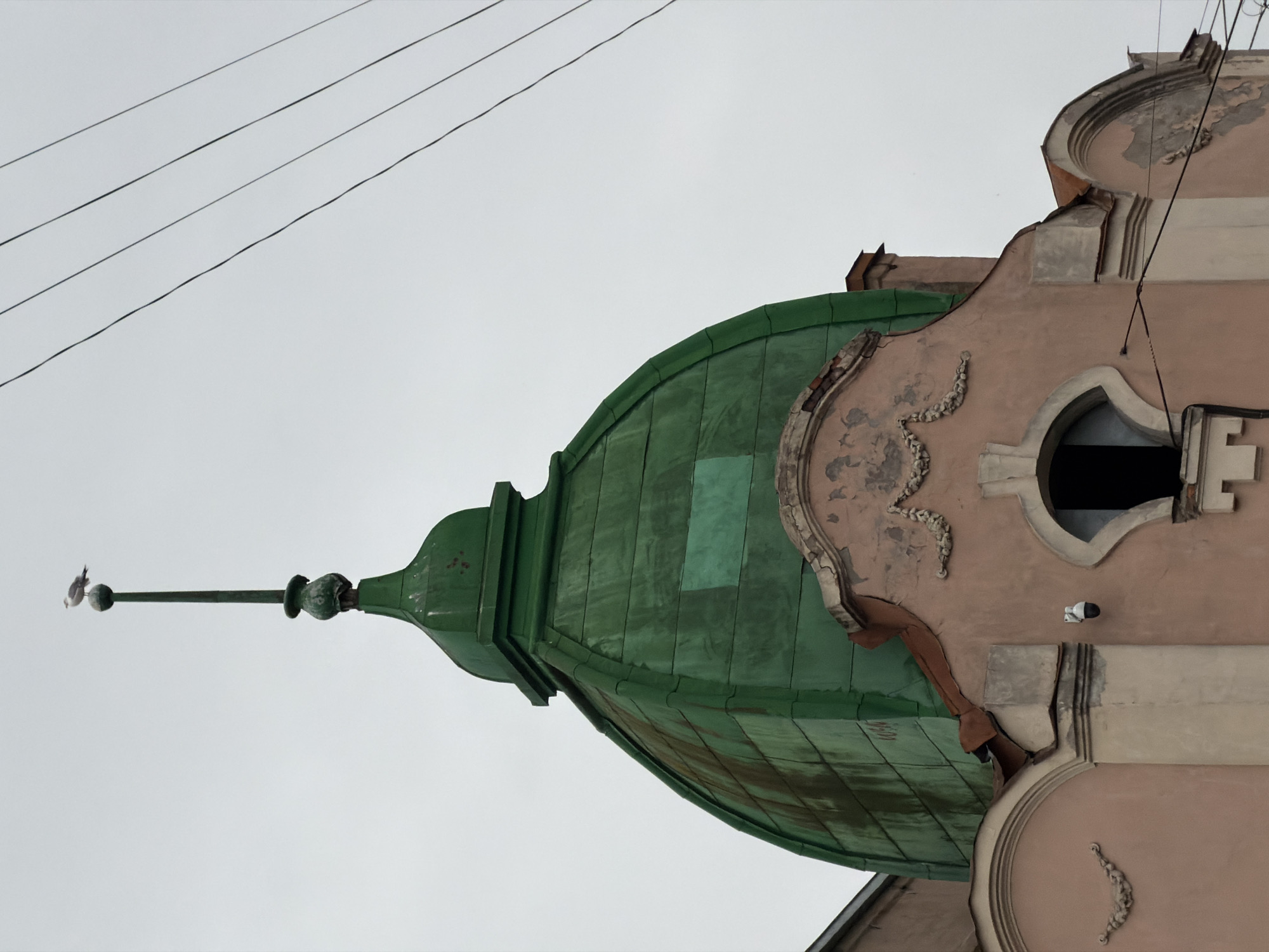 The image shows a section of an architectural structure, likely a building in Saint Petersburg, Russia, characterized by a green dome and ornate, light pink facade. The dome, made of what seems like metal or a similar material, features a pointed spire at its apex, topped with a perched bird. The facade is detailed with decorative elements, including baroque-style curves and embellishments around a small, dark window. Above, against a grey sky, electrical wires run parallel to the building. A security camera is mounted on the building near the window.