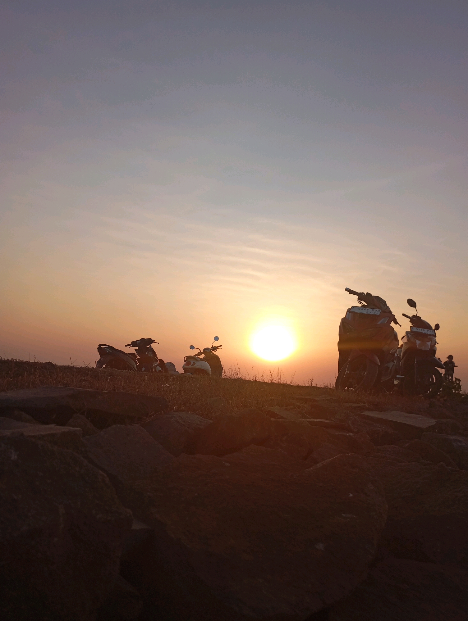 A low-angle shot captures a sunset over a rocky landscape with several parked motorcycles in the foreground. The sun, a bright orb, sits just above the horizon, casting a warm orange glow across the sky and illuminating thin, wispy clouds. The foreground consists of large, dark rocks, interspersed with dry grass and vegetation. Several motorcycles are parked on the elevated terrain behind the rocks, silhouetted against the bright sky. One white scooter is clearly visible on the right, while other dark motorcycles are parked to its left. In the far distance, on the right edge of the frame, a small, indistinct figure of a person is visible. The overall scene is serene and evokes a sense of tranquility. The image is believed to have been taken in Haurkolot, Indonesia.