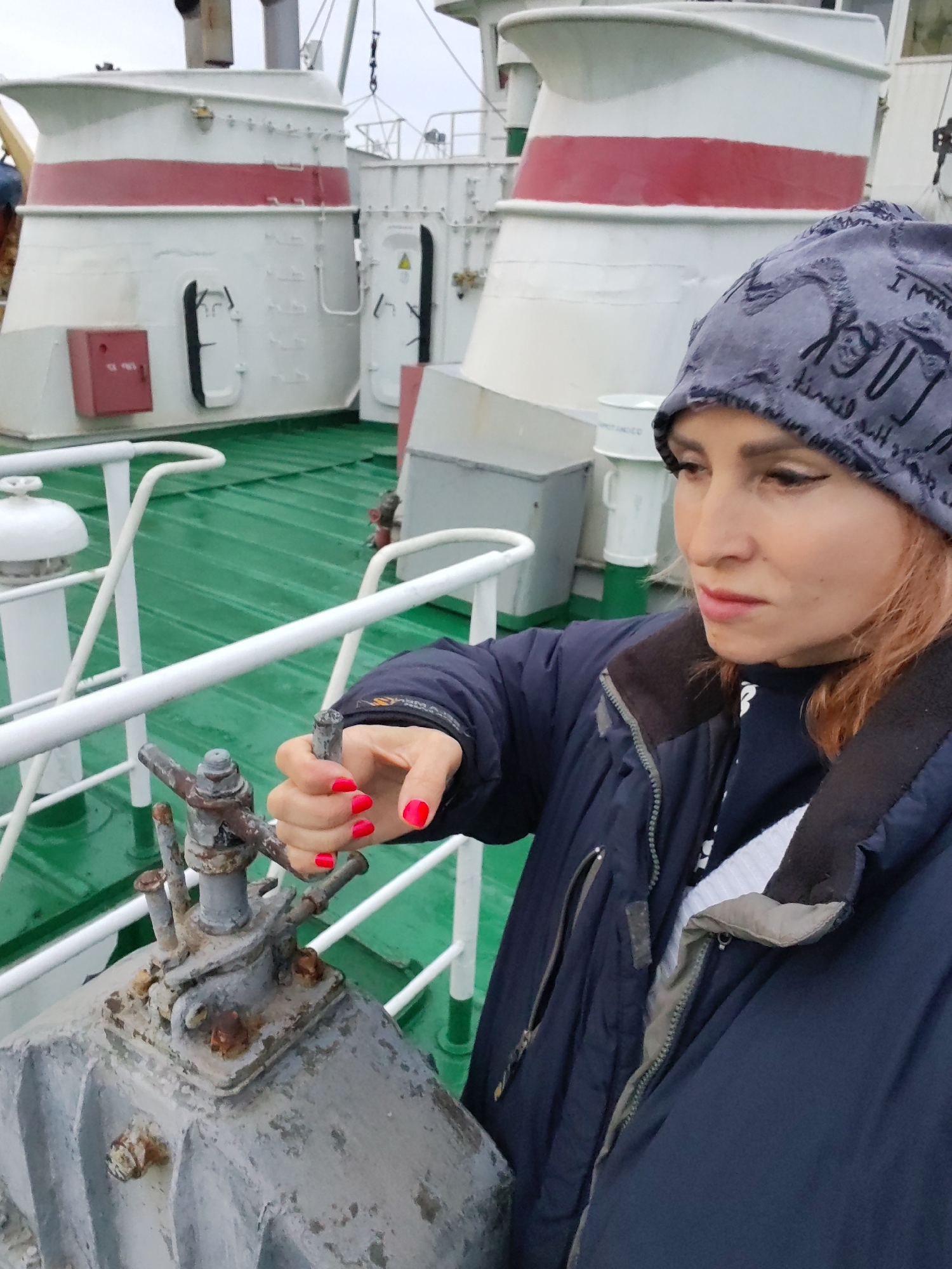 A woman with pink-dyed hair, wearing a dark blue jacket and a patterned beanie, is shown on the deck of a ship. She has bright red nail polish on her fingers and is using her right hand to operate a metal valve mechanism. The deck is painted green, and there are white railings and ship structures visible in the background, including a large white superstructure with a red stripe. The scene appears to be set outdoors in a cool or overcast environment. There is no information available to determine the specific location within Ivanov Bor, Russia.