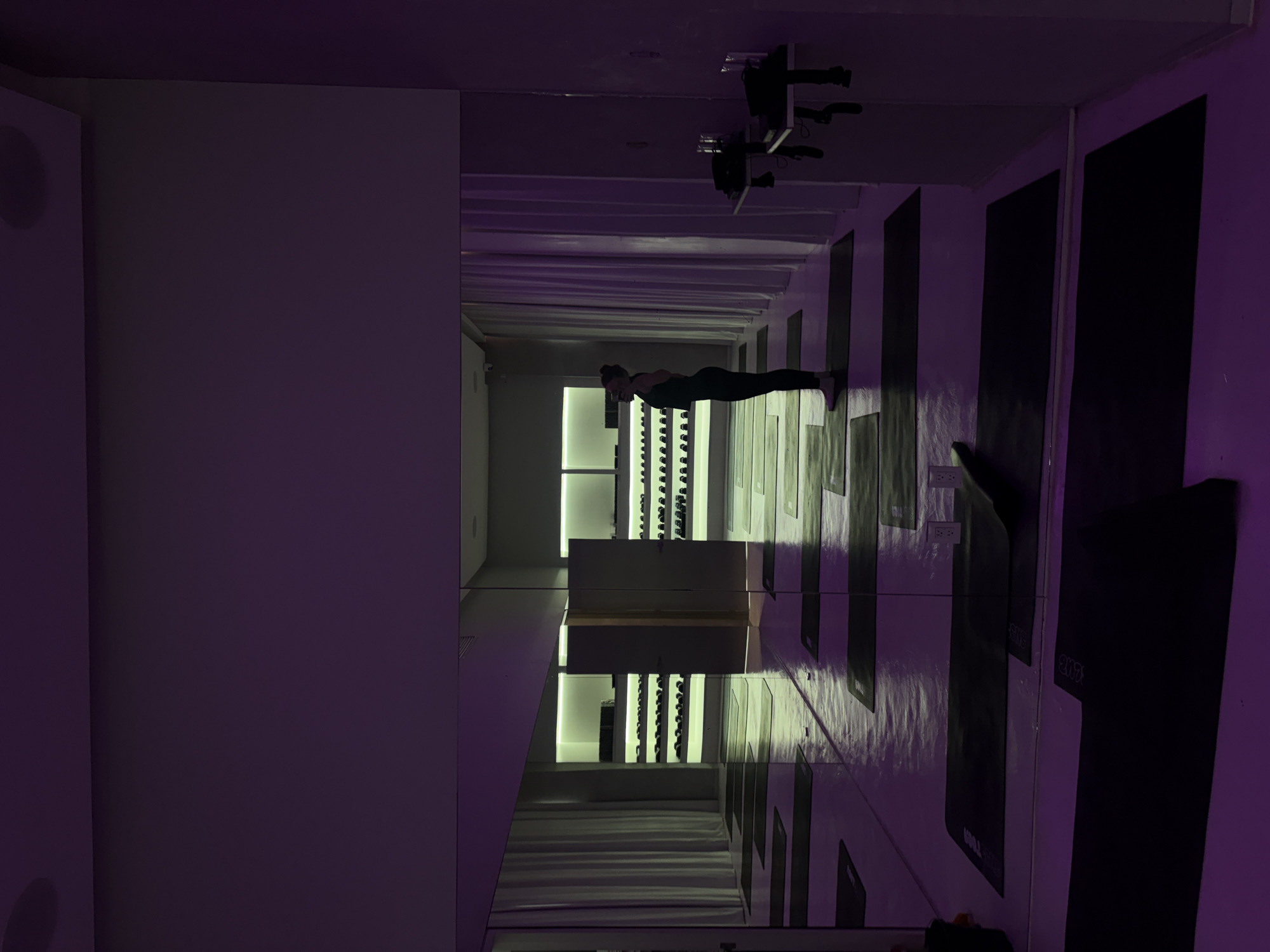 The image depicts a dimly lit yoga studio, bathed in purple ambient lighting. A woman, silhouetted and facing away from the camera, stands on a yoga mat in the center of the frame, her posture suggesting a yoga pose. The studio floor is a reflective surface, mirroring the yoga mats laid out in neat rows. To the left and in the reflection, white curtains are visible. In the background, shelves stocked with what appear to be water bottles are illuminated by white lights. The overall scene suggests a serene and organized space for physical practice. The image does not contain any information that would indicate a location in Mexico City, Mexico.