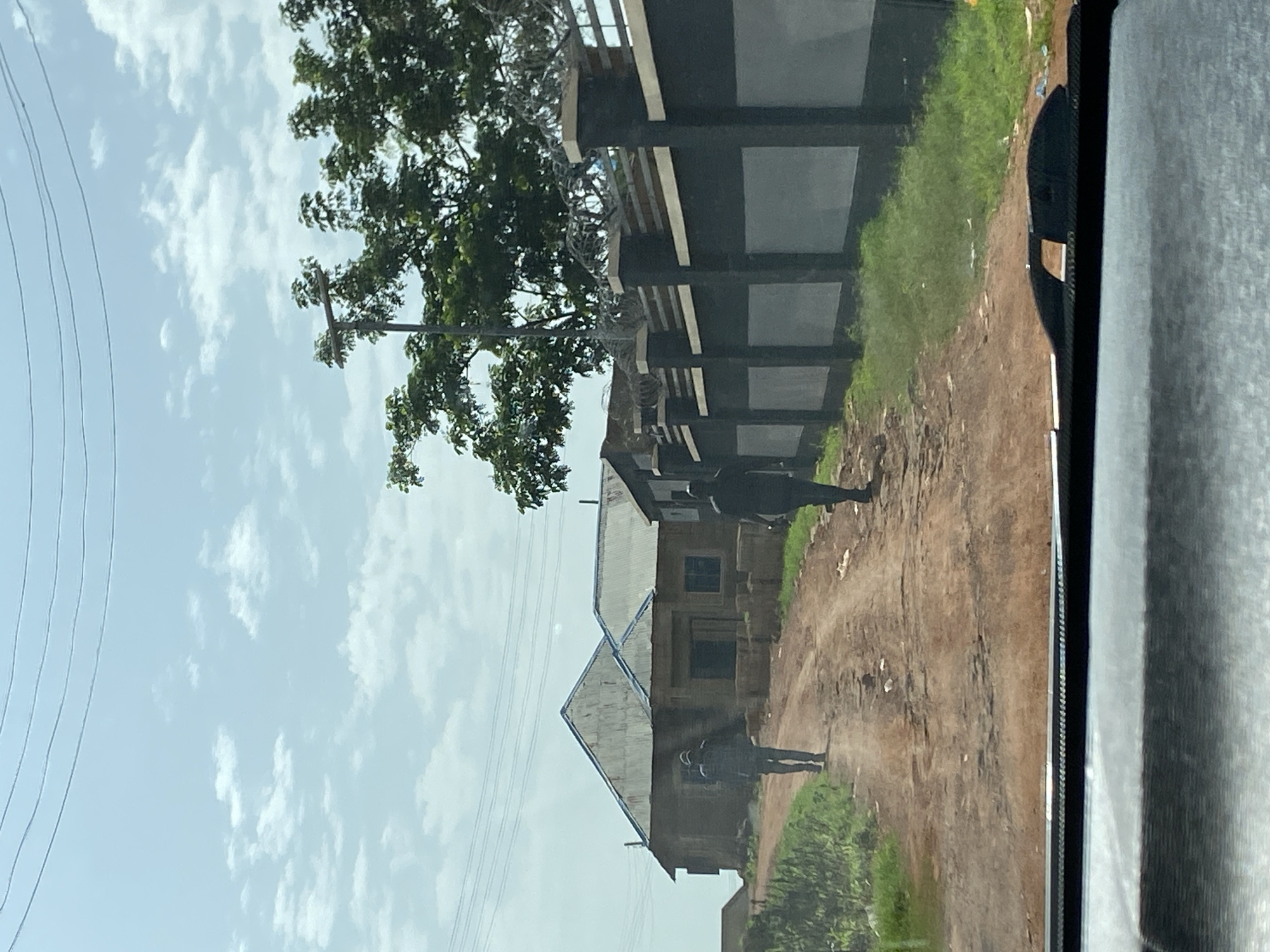 Two individuals are walking on a dirt road in Bar Jirgi Summa, Nigeria. The road is uneven with some debris. To the left of the road, there is a partially constructed building with a corrugated metal roof. To the right, there is a dark-colored fence with lighter panels and barbed wire at the top, backed by lush green vegetation and trees. The sky is partly cloudy. The foreground shows the dashboard of a vehicle.