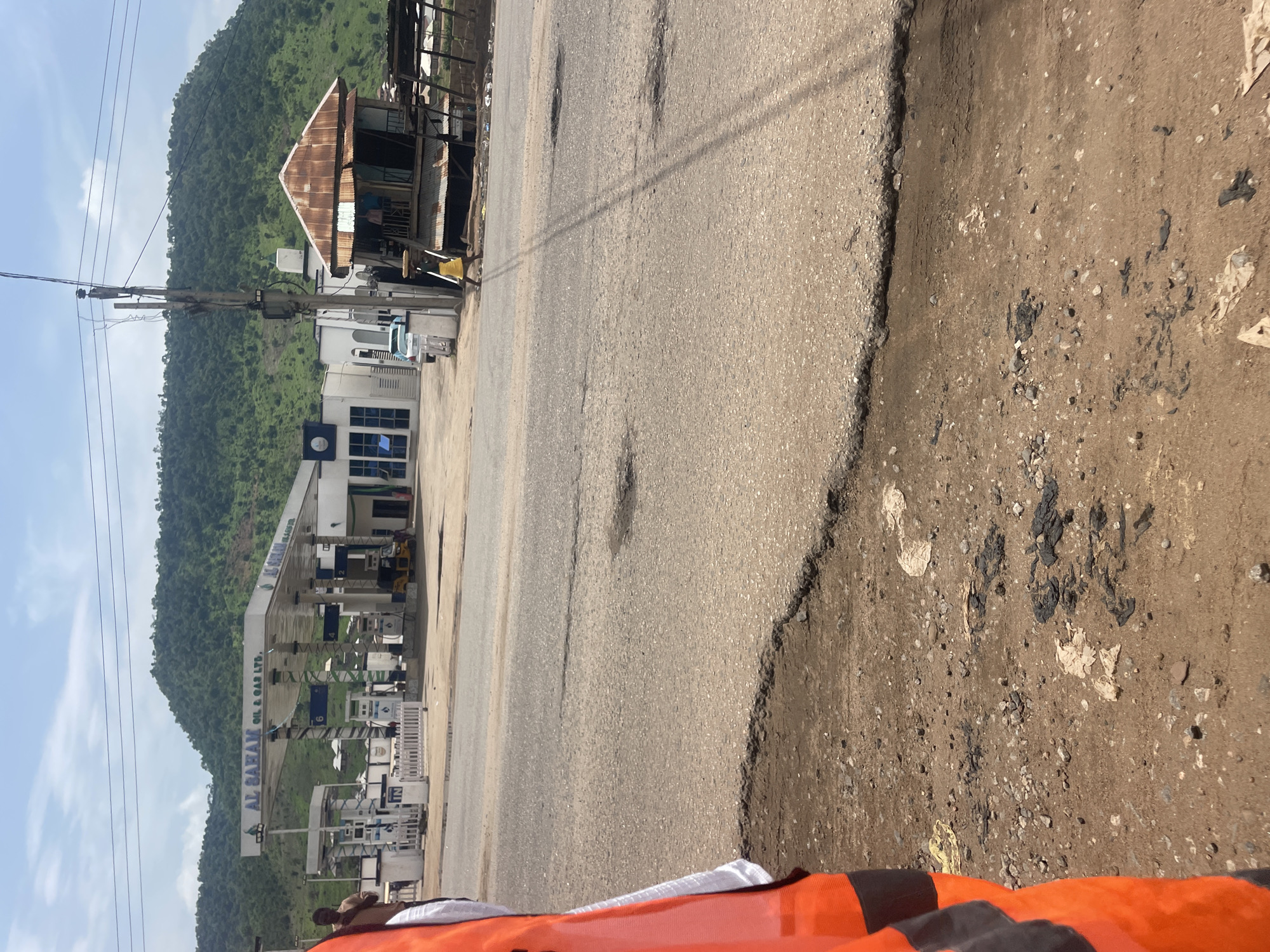 The image shows a street scene in Lokoja, Nigeria, with a gas station labeled "AL BAZAN OIL & GAS LTD." in the background. The gas station has multiple pumps and a modern building. To the right of the gas station, there is a rustic structure with a corrugated metal roof, appearing to be a small shop or dwelling. The street in the foreground shows signs of wear and tear, with potholes and damaged pavement. A person wearing an orange vest is partially visible on the left side of the frame. Power lines crisscross the sky. The overall environment suggests a semi-urban or roadside setting. A person in a yellow garment is walking on the road, carrying an object on their head.
