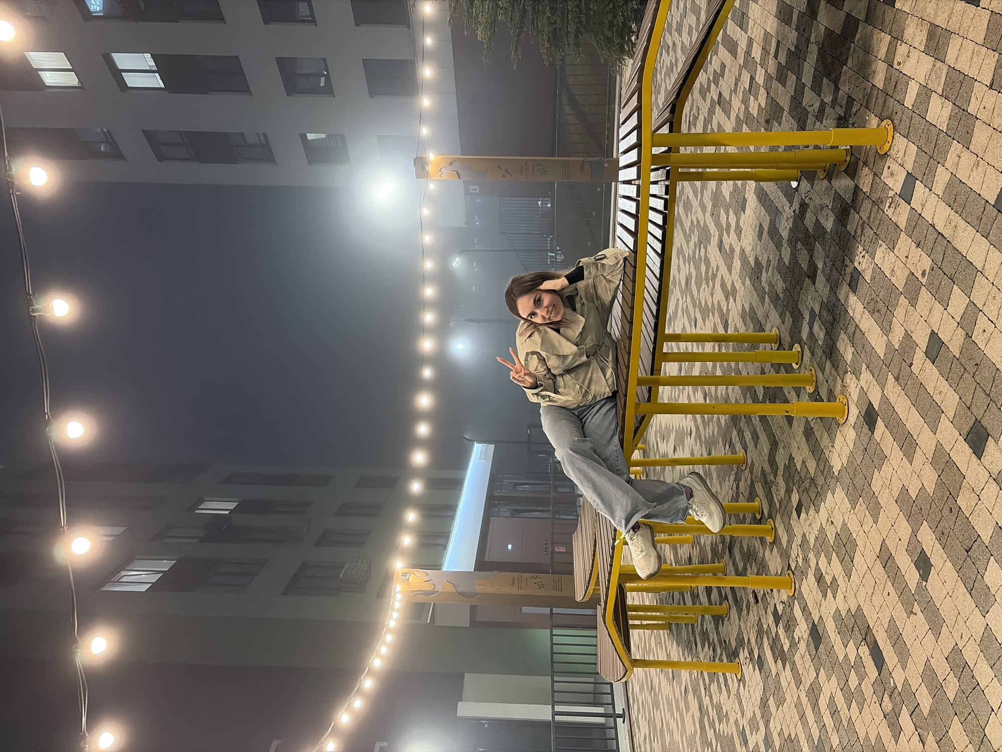 An eye-level, rotated photograph shows a young woman sitting on a yellow, tiered bench with her legs crossed, giving a peace sign with her right hand. She is wearing a light-colored jacket, jeans, and white sneakers. The bench is located on a patterned pavement made of square blocks of alternating gray and beige shades.
Behind the bench, there is a modern building with several windows, some of which are illuminated. A string of lights hangs above the bench and the building. The background is foggy, obscuring some details. Based on the available information, the location appears to be Izhevsk, Russia.