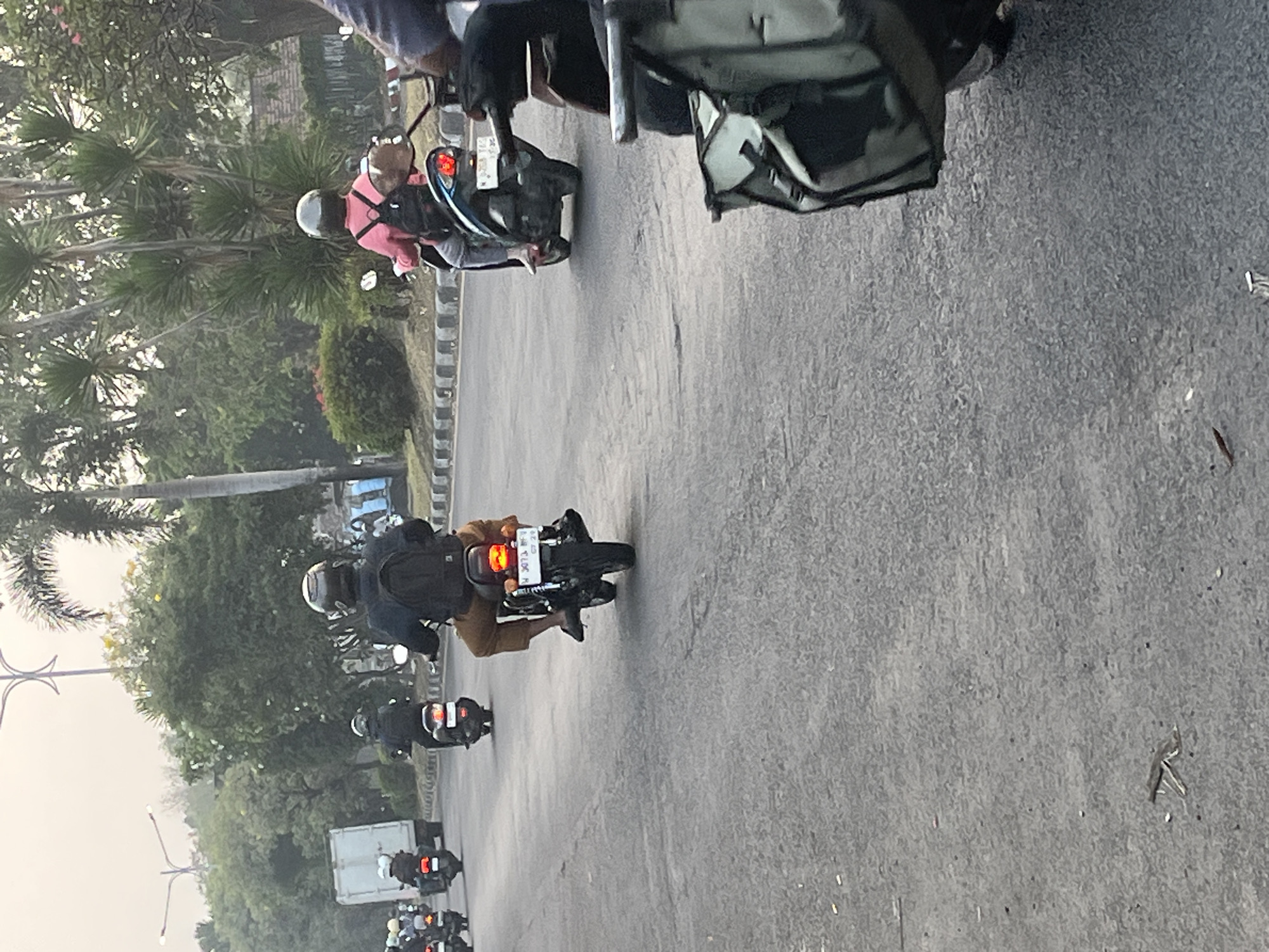 The image depicts a street scene in Kludan, Indonesia, with several individuals riding motorcycles. The road is paved and appears to be in moderate condition with some cracks. Trees and vegetation line the right side of the road, with a building visible in the background. The sky is overcast.
In the foreground, a motorcycle is partially visible on the right, with a person's arm and a black bag attached to the back of the motorcycle. Further down the road, a motorcyclist wearing a dark jacket and brown pants is riding a motorcycle. This rider has a black backpack. To the left of this motorcycle, another motorcyclist in a black jacket and helmet is riding, also with a backpack. In the distance, several other motorcycles are visible, including one with a rider in a pink shirt and a backpack. A white truck is also present in the far left background. The overall impression is a typical street scene with motorcycle traffic in Indonesia.