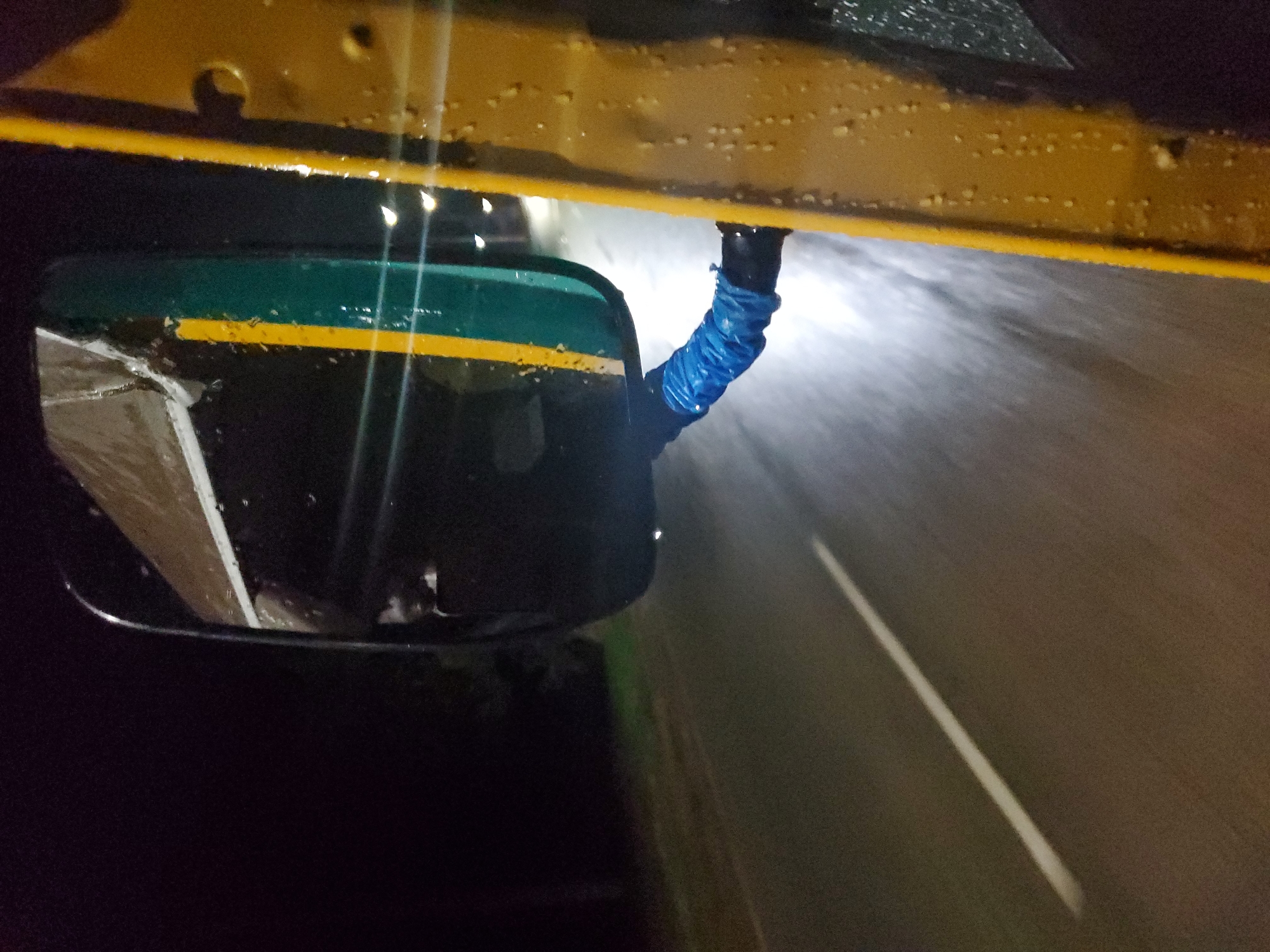 The image depicts a nighttime or low-light scene from the perspective of a vehicle, possibly in Vom, Nigeria. The foreground shows a section of a yellow vehicle body, likely a car or bus, speckled with water droplets, suggesting rain or wet conditions. A side mirror is prominently featured on the left, which is dark green with a bright horizontal yellow stripe. The arm connecting the mirror to the vehicle is covered in a textured, crinkled blue material, possibly a protective sleeve.
The reflection in the side mirror is dark and indistinct, showing what appears to be part of the vehicle's interior or another structure. Beyond the vehicle, a dark, wet road surface is visible, illuminated by what appears to be a headlight beam, creating a bright, blurred streak of light on the right side of the image, implying movement. A clear white lane marking runs diagonally across the dark asphalt in the lower right portion of the frame. There is no visible text in the image. The overall impression is of driving on a wet road during the night.