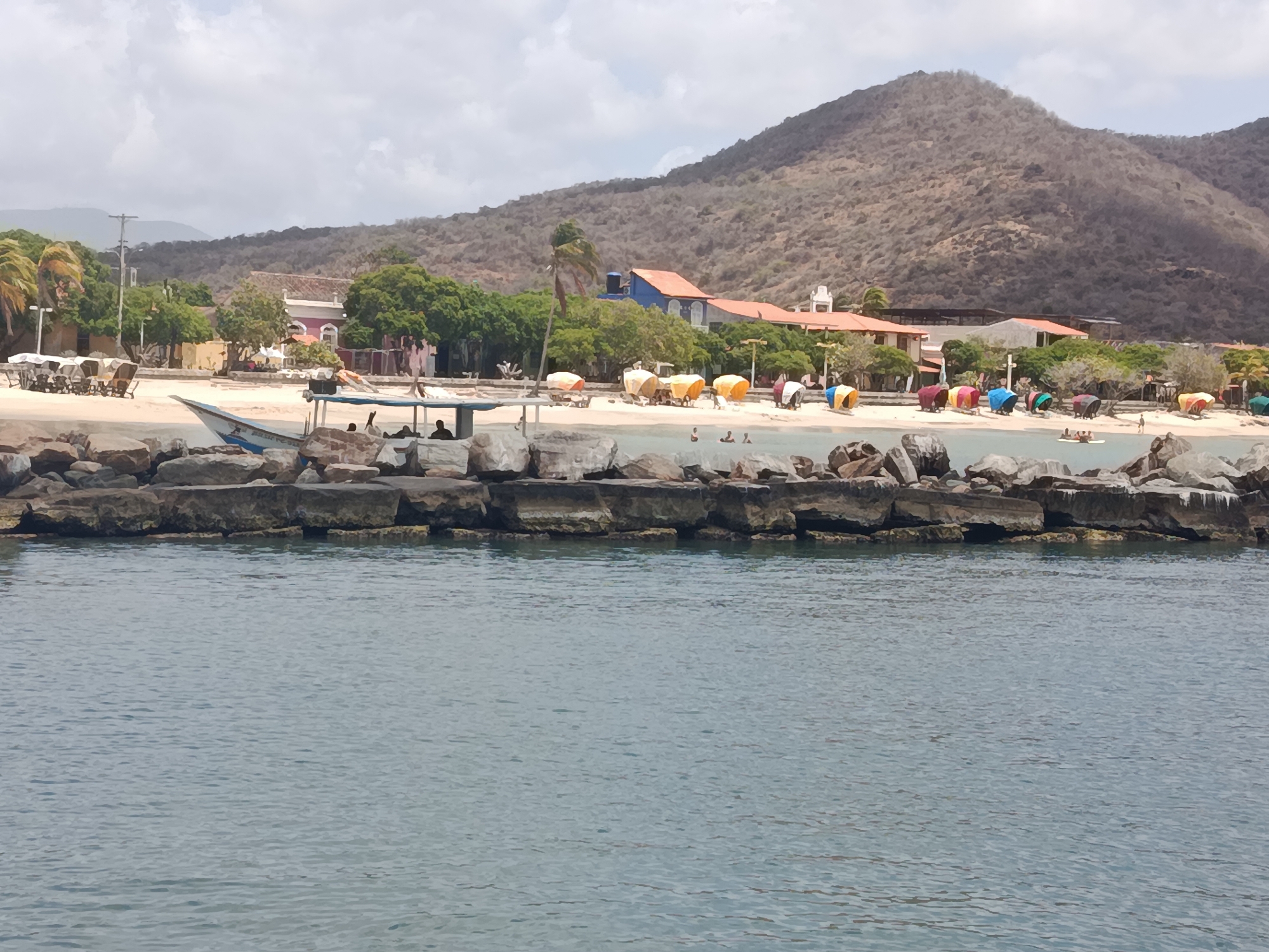 The photograph depicts a coastal scene in Pedro González, Venezuela. In the foreground, calm, grey-blue water stretches across the majority of the image. A barrier of large, grey-brown rocks extends horizontally across the middle ground, separating the water from a sandy beach.
On the beach, numerous colorful beach umbrellas and various buildings are apparent. The buildings have red-tiled roofs and white walls. The backdrop is dominated by a large, barren mountain. The sky is overcast with grey and white clouds. Several people are visible on the beach. A boat with a canopy is also visible near the rocks.