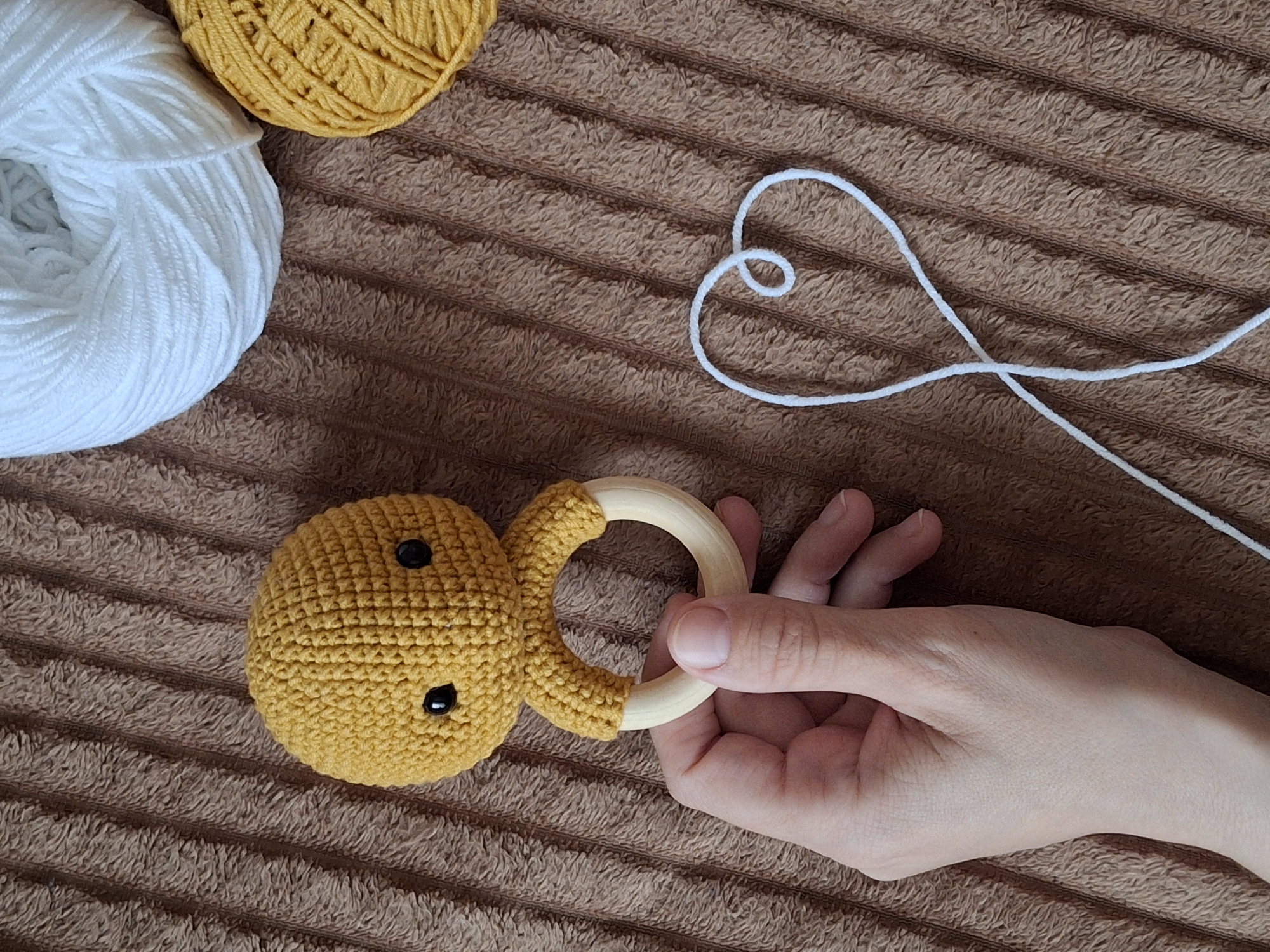 A high-angle shot shows a brown textured surface, potentially a blanket. Located in the upper left corner are two balls of yarn, one white and one yellow. To the right of the yarn, a length of white yarn forms a heart shape. Near the bottom left corner, a yellow crocheted object resembling a smiling face is visible with two black button eyes. A wooden ring is attached to the crocheted object. A hand grips the wooden ring, holding the toy. The photograph was taken in the city of Chaikovsky, Russia.