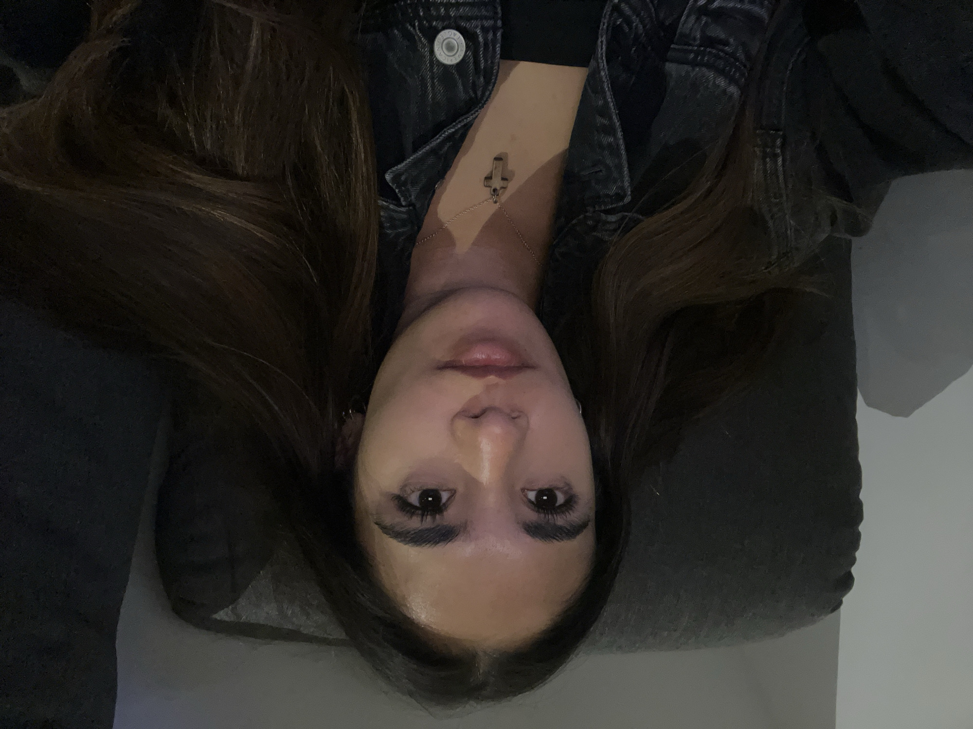 The image shows a woman lying down, with her head facing the camera and her hair spread around her head. She is wearing a dark denim jacket and a necklace with a cross pendant. She is resting on what appears to be a gray cushion. The background is indistinct, with some possible wall space visible on the right. The image does not provide enough information to identify a specific location in San Bernardino Tlaxcalancingo, Mexico.