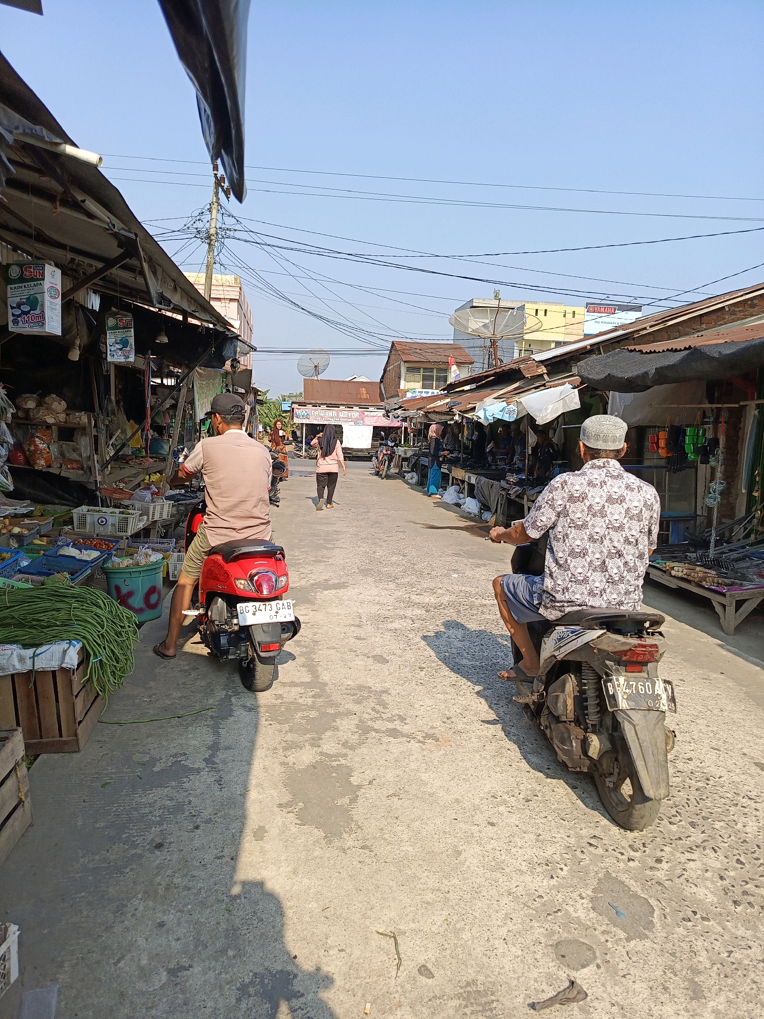 The image depicts a street scene in the city of Megang Sakti I, Indonesia. The street is lined with market stalls on both sides, covered by corrugated metal roofs and tarps, suggesting an outdoor marketplace. In the foreground, two men are riding motorbikes. The man on the left is on a red scooter, wearing a light brown t-shirt, olive shorts, and a black baseball cap. The man on the right is on a white and black scooter, wearing a patterned shirt, blue shorts, and a white patterned cap. The street is paved with concrete. Various goods are visible in the market stalls, including produce like long beans and tomatoes, and various packaged items. Power lines crisscross overhead. The sky is clear and blue, indicating a sunny day. In the background, other people are visible walking and riding motorbikes, and some buildings are present. The overall impression is a busy local market area on a clear day.
