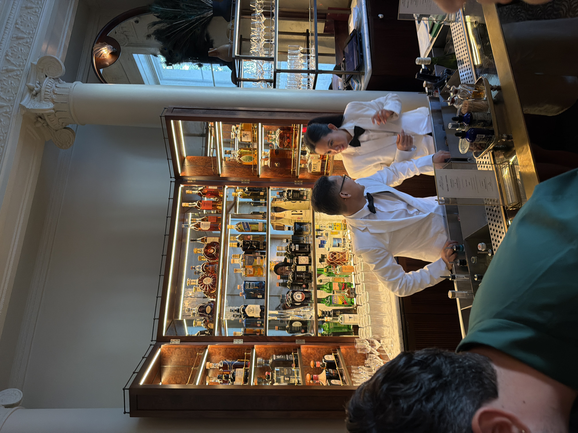 A medium shot captures a bar scene with two bartenders dressed in white suits and bow ties. The bar is stocked with various bottles of liquor on illuminated shelves, featuring brands like Knob Creek and Hennessy. The backdrop includes classical architectural elements like columns and ornate moldings. In the foreground, a person wearing a green shirt is partially visible, facing away from the camera, appearing to interact with the bar. The overall setting suggests a sophisticated establishment, potentially in London, United Kingdom, given the visual cues of refined decor.
