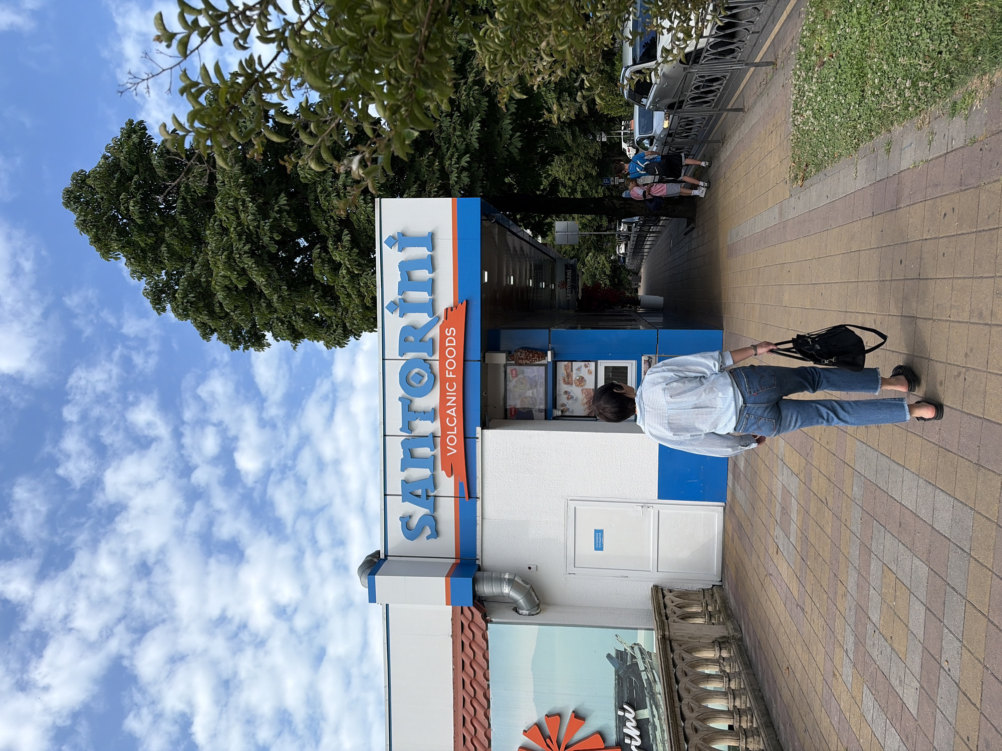 The image shows a street scene in Sochi, Russia. In the foreground, a person walks away from the camera on a tiled sidewalk. The person is wearing a light blue striped shirt, blue jeans, and black sandals, and carrying a black tote bag. To the left of the sidewalk is a white building with blue accents, a large sign that reads "SANTORINI VOLCANIC FOODS", and a mural of a windmill and boat. To the right of the sidewalk is a road with parked cars and a metal fence. In the background, there are lush green trees and a cloudy blue sky. The overall impression is a bright, sunny day in a city setting.