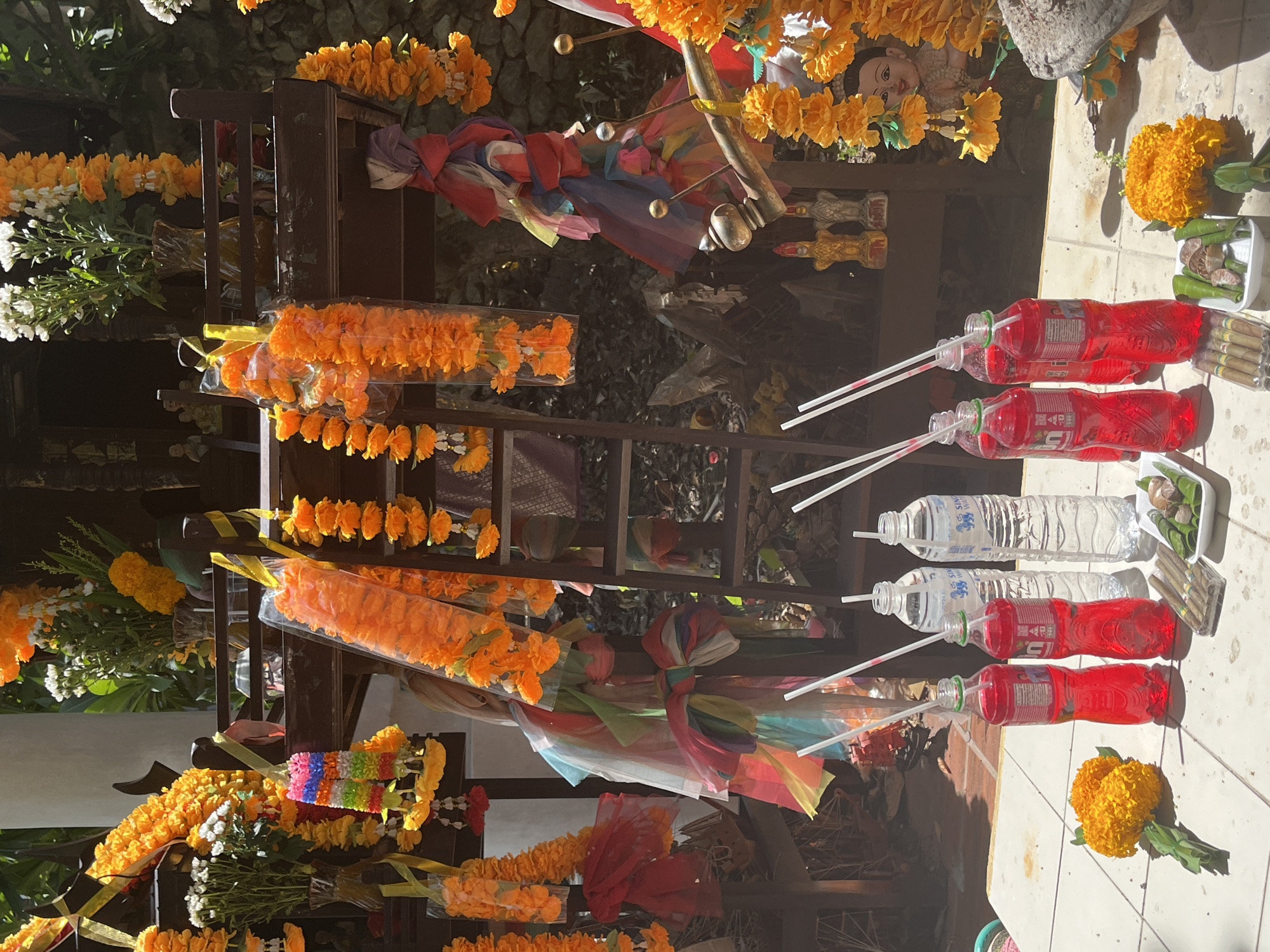 A photograph taken outdoors in Baan Hua Thanon, Thailand, depicts a shrine or altar. Garlands of orange and yellow flowers are draped across the wooden structure of the altar. Brightly colored fabrics are tied in bows. Bottles of water and red soda with straws inserted are placed on the floor, alongside marigold flowers, betel leaves, and small cigars. The scene is lit by sunlight.